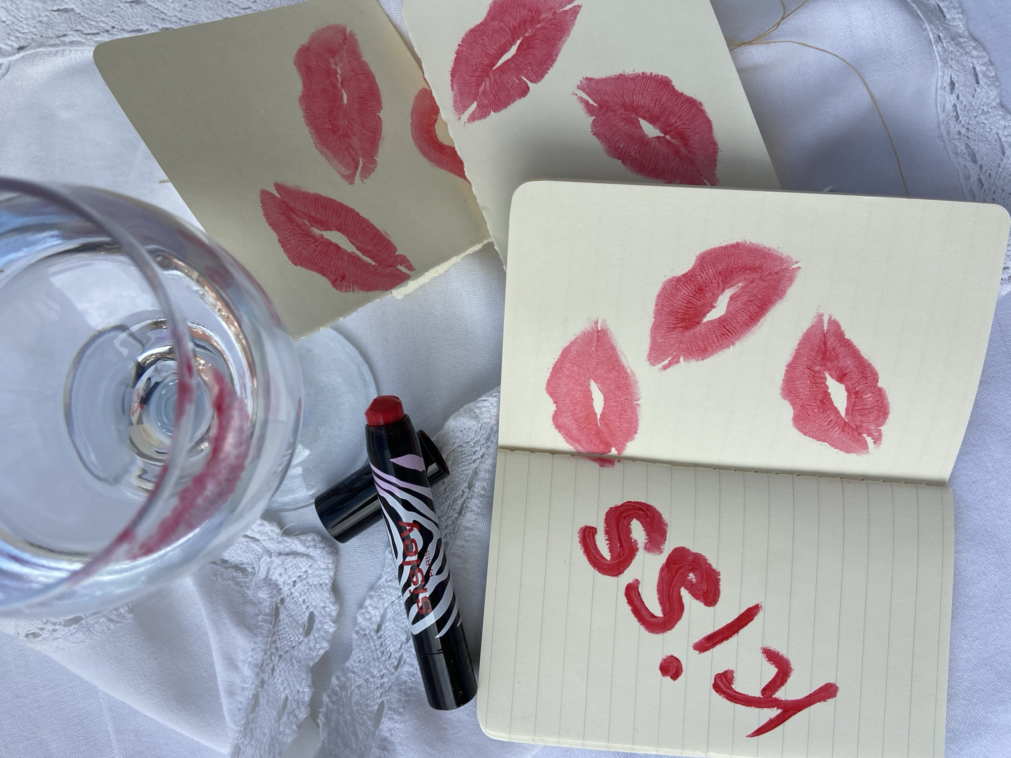The image shows a still life scene on a white lace-trimmed cloth. In the lower left, there's a clear wine glass with lipstick smudges around the rim, positioned slightly askew. Adjacent to the glass, to its right, is a black lipstick tube with a zebra stripe design and a red cap, lying horizontally. There are three off-white paper cards, each bearing multiple lipstick kiss prints, scattered above the glass and lipstick. The top card has three kiss marks, the one to its right has two, and the card at the bottom, is open and has two kiss marks. On the open card, the word "Kiss" is written in red lipstick. The scene is set in an unknown location and there is no indication that the image was taken in Budva, Montenegro.