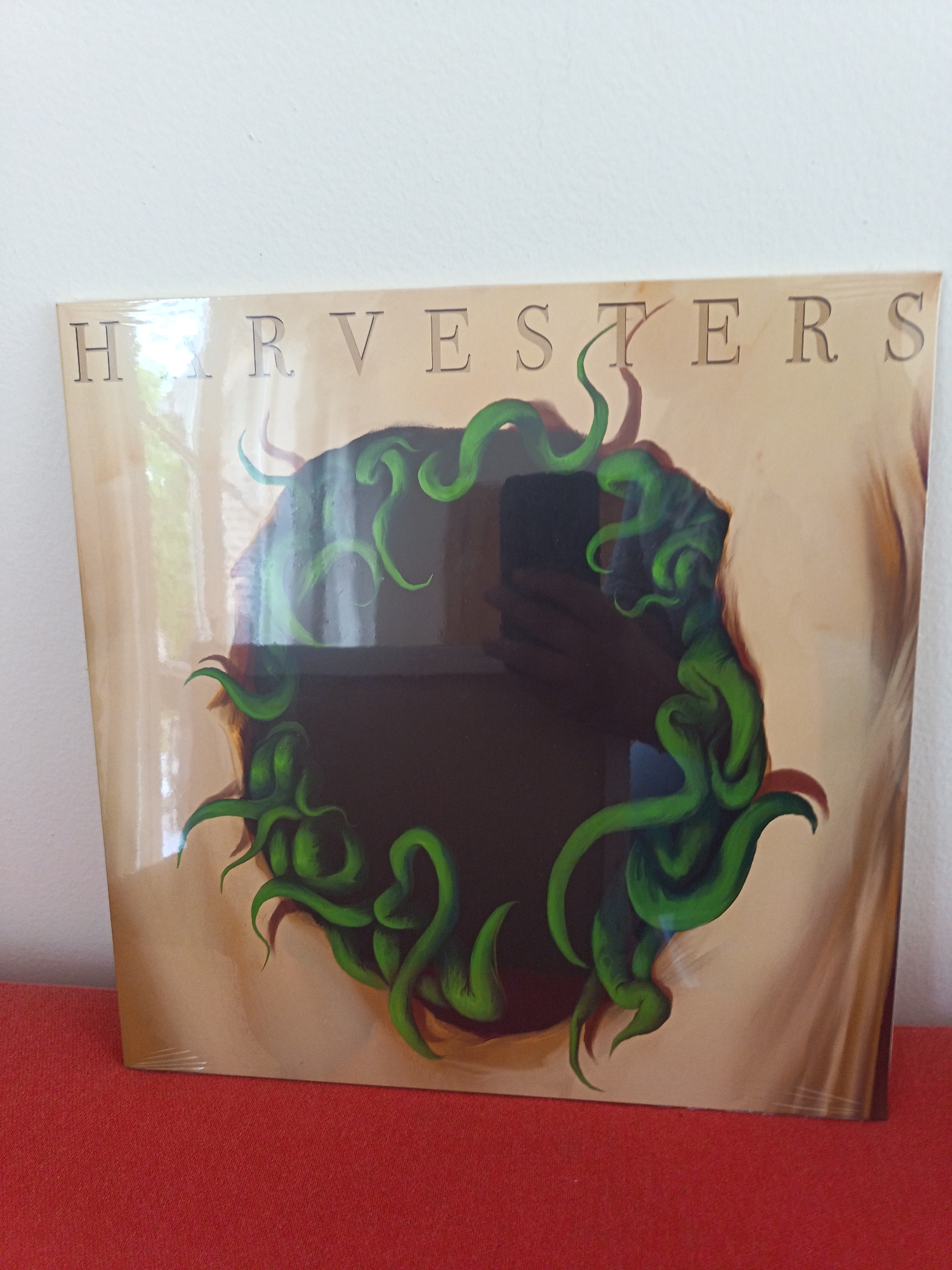 The image shows a vinyl record album titled "HARVESTERS". The album cover features a tan-colored background with a large, dark, circular void in the center. Green, tentacle-like forms surround the void. The album appears to be in a protective plastic sleeve and is resting on a red surface. The background behind the album is a white wall. There is no indication of a city or specific location.