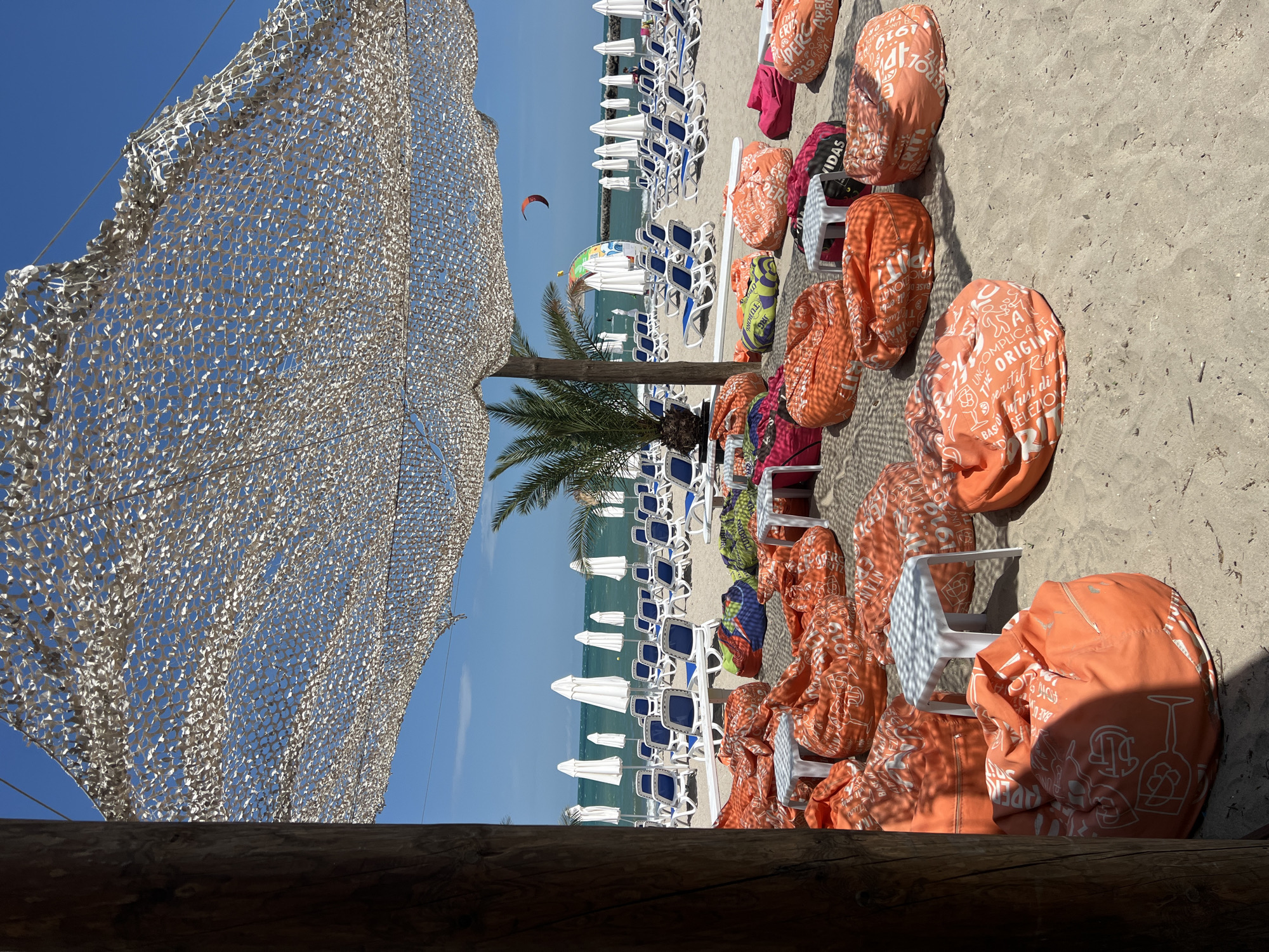 This is a photograph taken on the beach in Pomorie, Bulgaria, likely in the summer. The foreground is dominated by numerous large, orange beanbag chairs scattered across the sand. Some of these beanbag chairs have white lettering and graphics printed on them. Small, white square tables are interspersed among the beanbag chairs. A portion of a wooden post is visible on the left side of the image, suggesting a structure or part of a beach setup.
In the mid-ground, rows of white beach umbrellas are neatly arranged behind white sun loungers with blue seating. The sea is visible in the background, under a clear blue sky. A small palm tree is situated behind some of the beanbag chairs, adding to the tropical beach atmosphere. A kite in the sky and a colorful archway can also be seen near the water's edge. The overall scene depicts a relaxed and inviting beach setting, likely a designated area for patrons of a beach bar or resort.