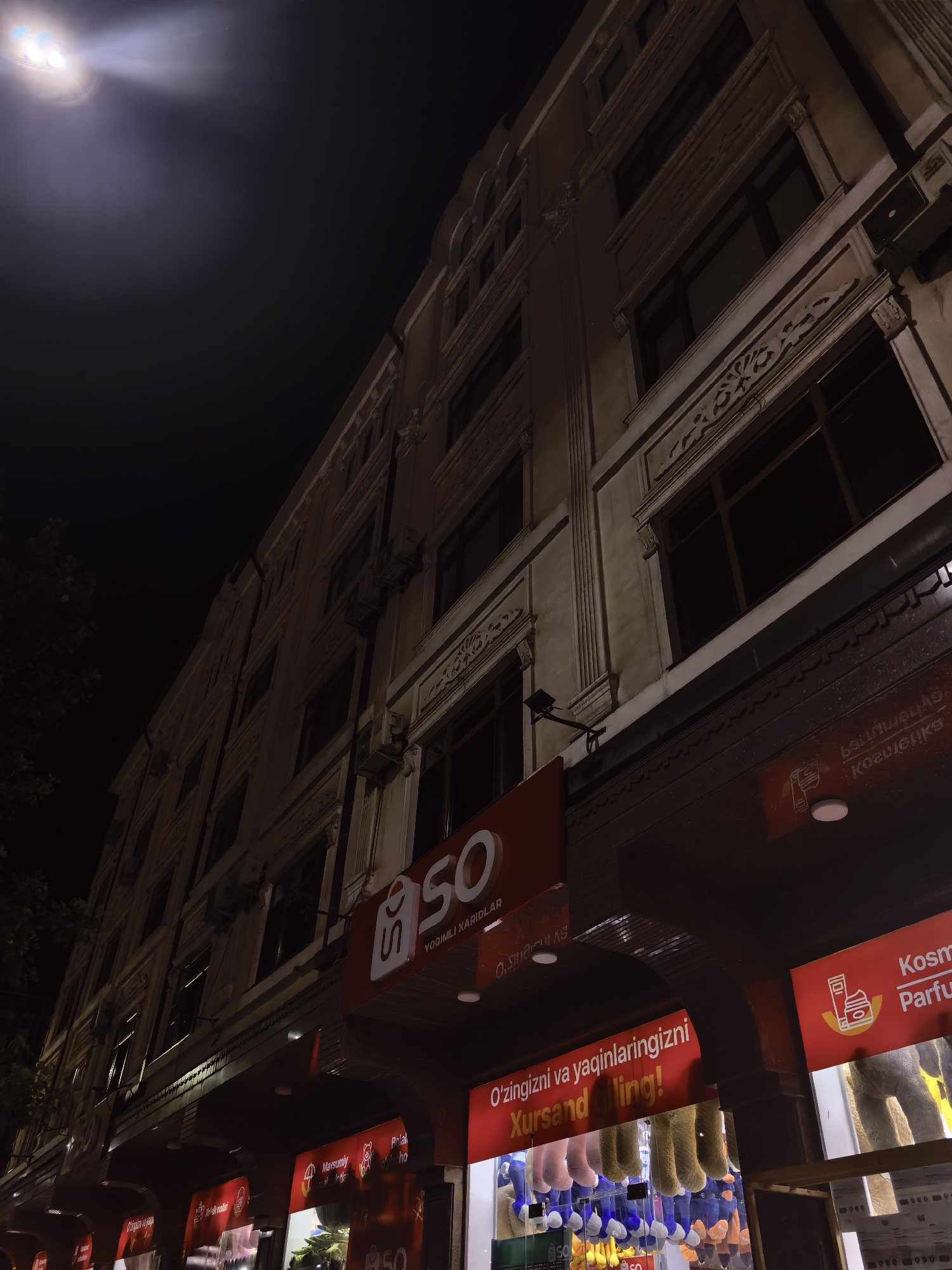 The image captures a nighttime street view in Fergana City, Uzbekistan, focusing on the facade of a multi-story building with shops at its ground level. A bright, diffused light source, possibly a large street lamp or spotlight, illuminates the upper portion of the scene from the left, casting a significant glow. The building itself appears to be of classic architecture, with decorative detailing around the windows and along the facade.
Below the main facade, several shop signs are visible, predominantly in red with white and black text, suggesting retail establishments. One prominent sign reads "SO" in large white lettering on a red background, with smaller text below. Another sign displays "Kosmetika Parfume" indicating cosmetic and perfume products. Beneath these signs, a display of stuffed animals, primarily teddy bears in various colors, is visible through shop windows, suggesting a toy or gift shop. The overall impression is of a commercial street at night, with shops open for business. The surrounding environment is dark, highlighting the artificial lighting from the shops and the street lamp.