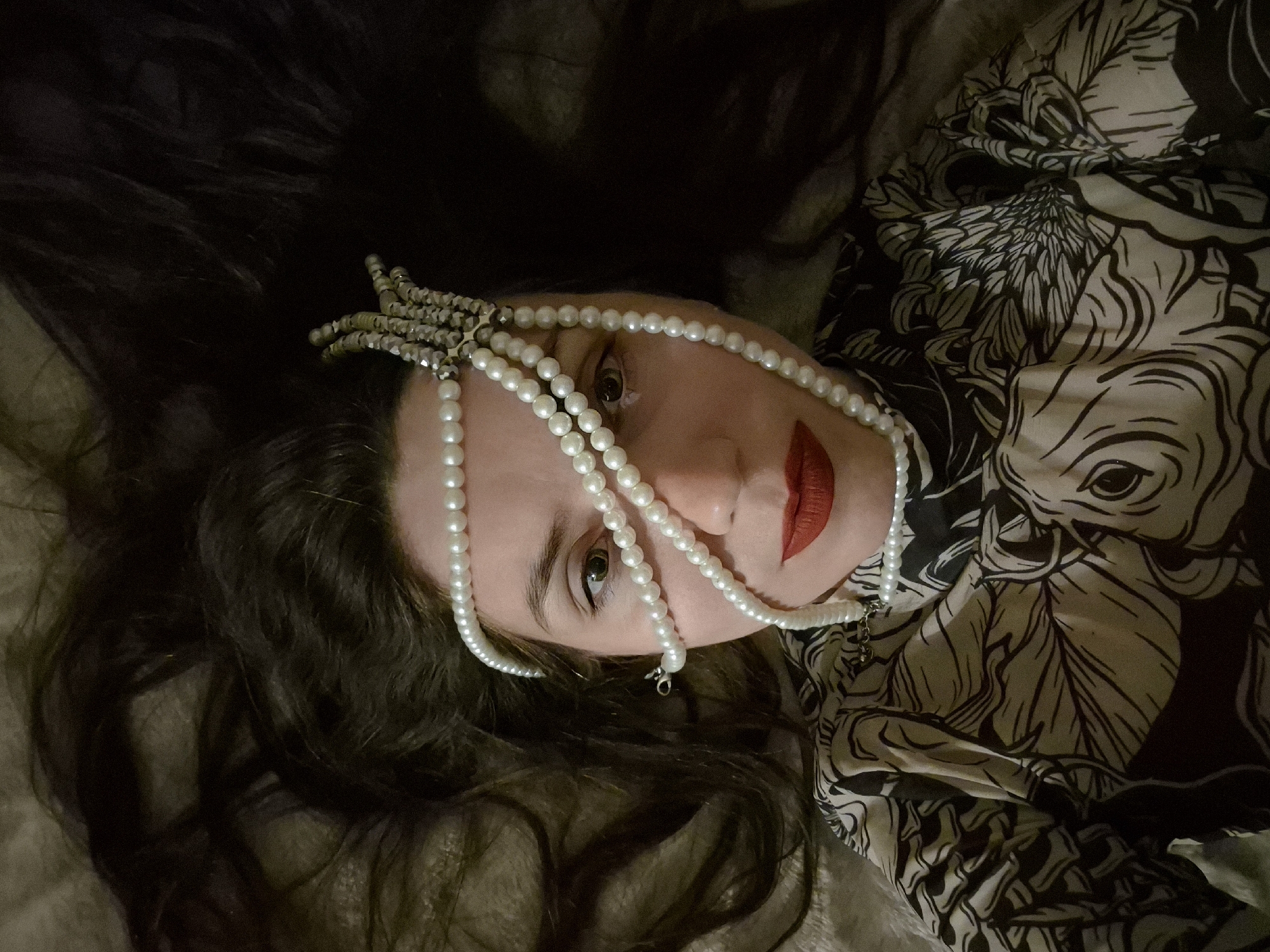 The image is a close-up, overhead shot of a woman lying on a bed with her long, dark hair spread out. She is wearing a black and white patterned garment and a pearl necklace is draped across her face, obscuring parts of her eyes and forehead. Her lips are painted with red lipstick. The background appears to be a textured, neutral-colored surface, likely bedding. There are no discernible elements that would indicate the location within the city of Chaikovsky, Russia.