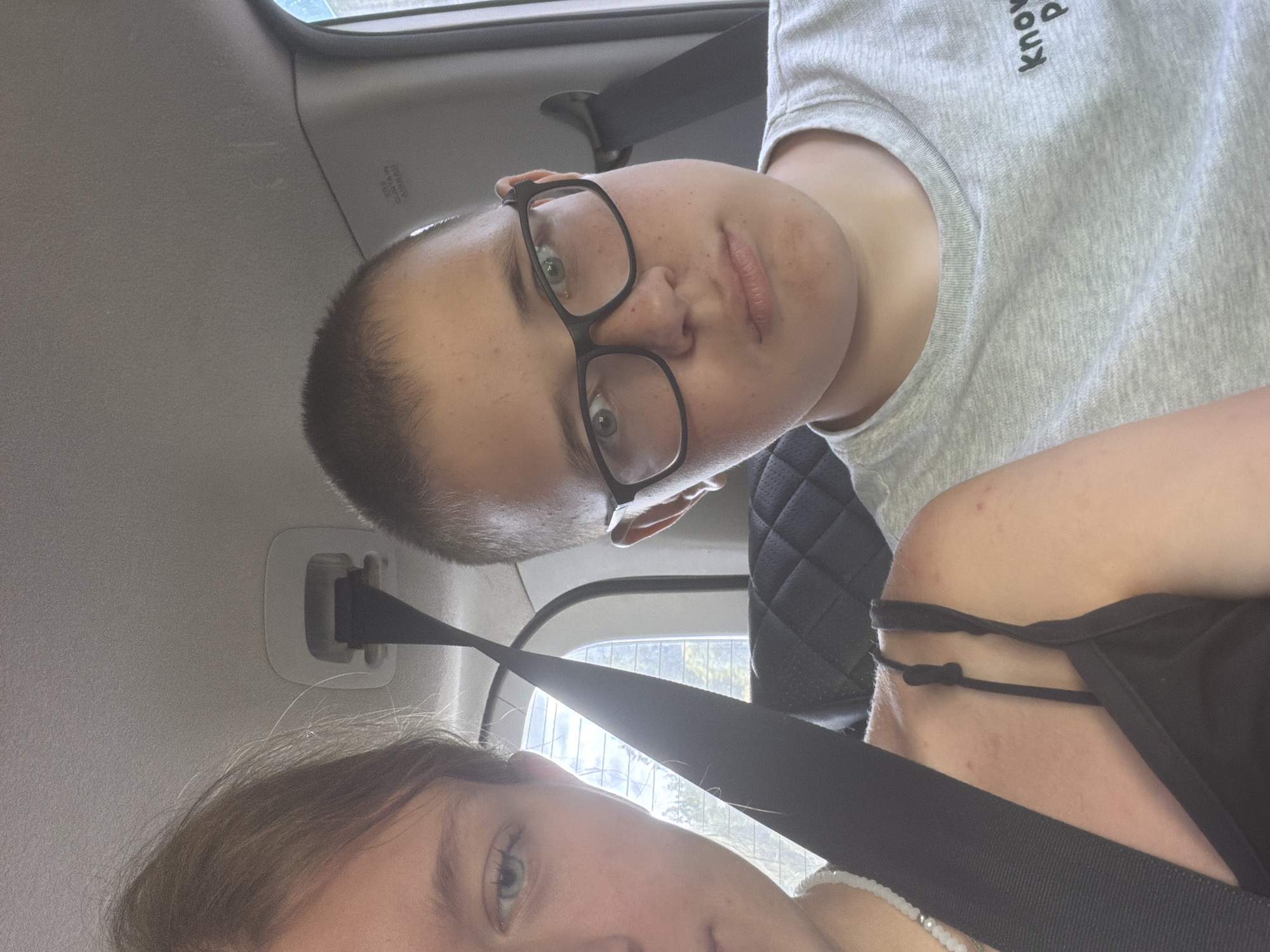 Two individuals, a female on the left and a male on the right, are seated in the back of a vehicle, with a seatbelt crossing between them. The female's left side of her face and shoulder are visible, and she is wearing a black tank top and a beaded necklace. The male is wearing glasses and a grey t-shirt. The background shows the interior of the car, including the car's ceiling and a window with trees visible outside. There is no indication of the specific city of Novaya Zhizn', Russia.