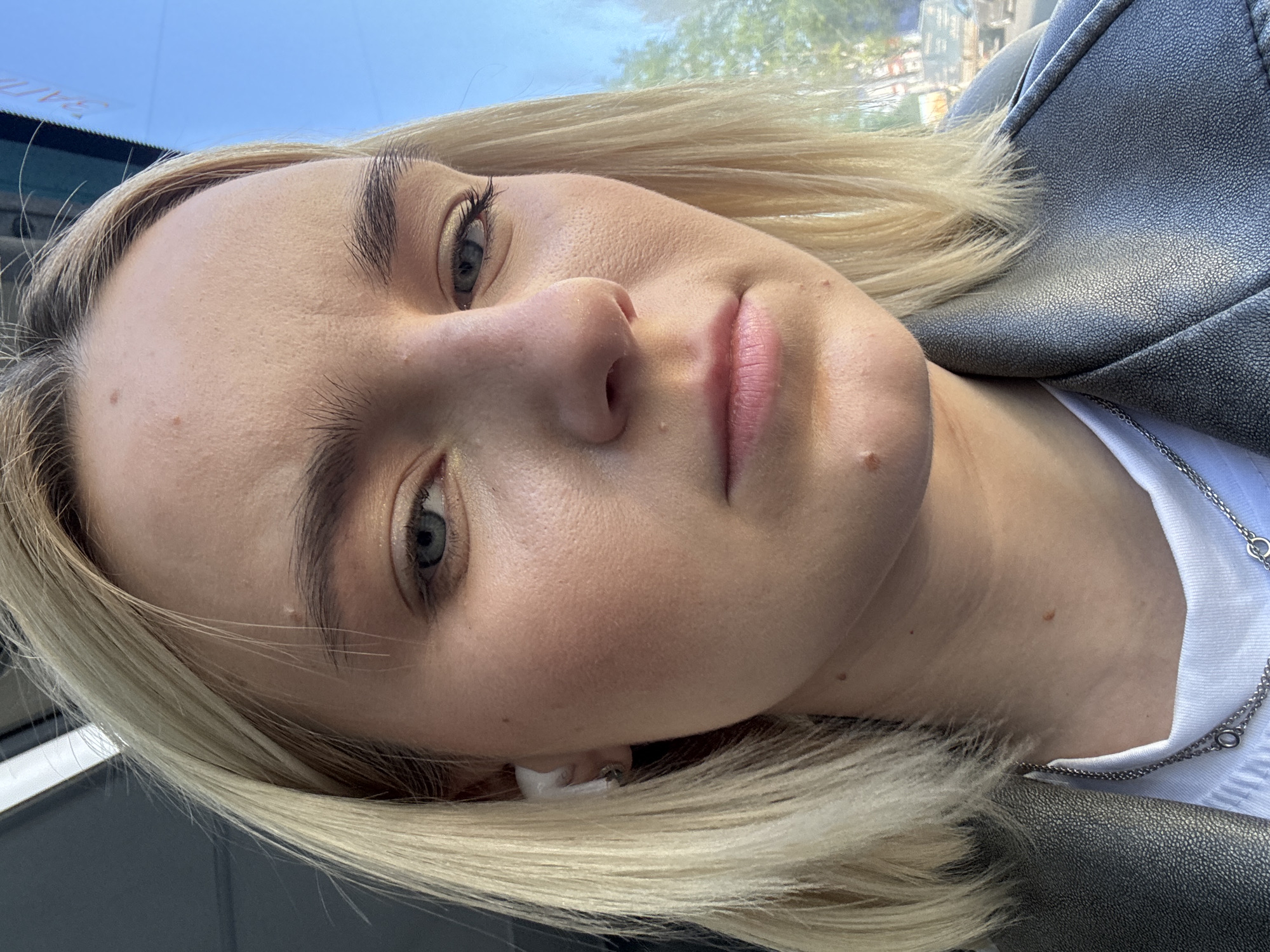 A close-up, angled portrait shows a fair-skinned woman with short, blonde hair. Her eyes are light-colored. She has a mole on her cheek. She is wearing an earring in her ear and is attired in a white shirt underneath a gray leather jacket, with a silver chain necklace visible. The background includes an indiscernible city view, partially visible through a window. Based on the urban architecture and potential landmarks, the location may be the city of Saint Petersburg, Russia.