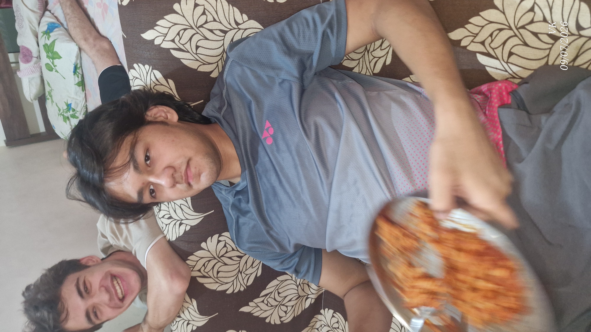 In a room in Vrindavan, India, two young men are seen sitting on a sofa. One man in the foreground, wearing a grey t-shirt, is holding a plate of food. The other man, in the background, is leaning over the back of the sofa and laughing. The sofa is covered with a patterned fabric. A bed is visible behind the men, with pillows and a person's legs resting on it. The room appears to be a living space or bedroom.