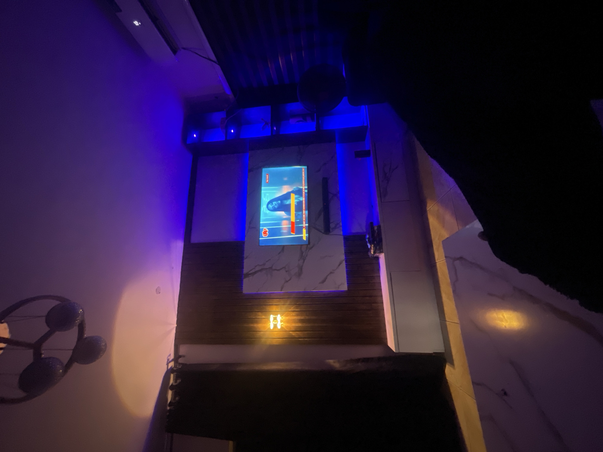 The image depicts an interior space, likely a room, with a modern or contemporary design. The color palette is dominated by shades of blue and purple lighting. The focal point is a centrally mounted digital display showing a car racing game interface. The wall behind the display features a mix of materials: sections of white marble-like paneling alternating with dark wood panels. A decorative light fixture sits below the central display on the wooden section of the wall. The bottom of the image is obscured by what appears to be a dark-colored couch or seating area. In the left foreground is a decorative wall-mounted light fixture, possibly a sculptural piece. The right portion of the image is dominated by a large dark mass, likely an object close to the lens or a person standing in the way of the camera's view. The room is illuminated with ambient blue lighting, with some directional lighting illuminating the features on the wall. There is no specific indication of this image being from Lokoja, Nigeria. The scene lacks any people or interactive elements.