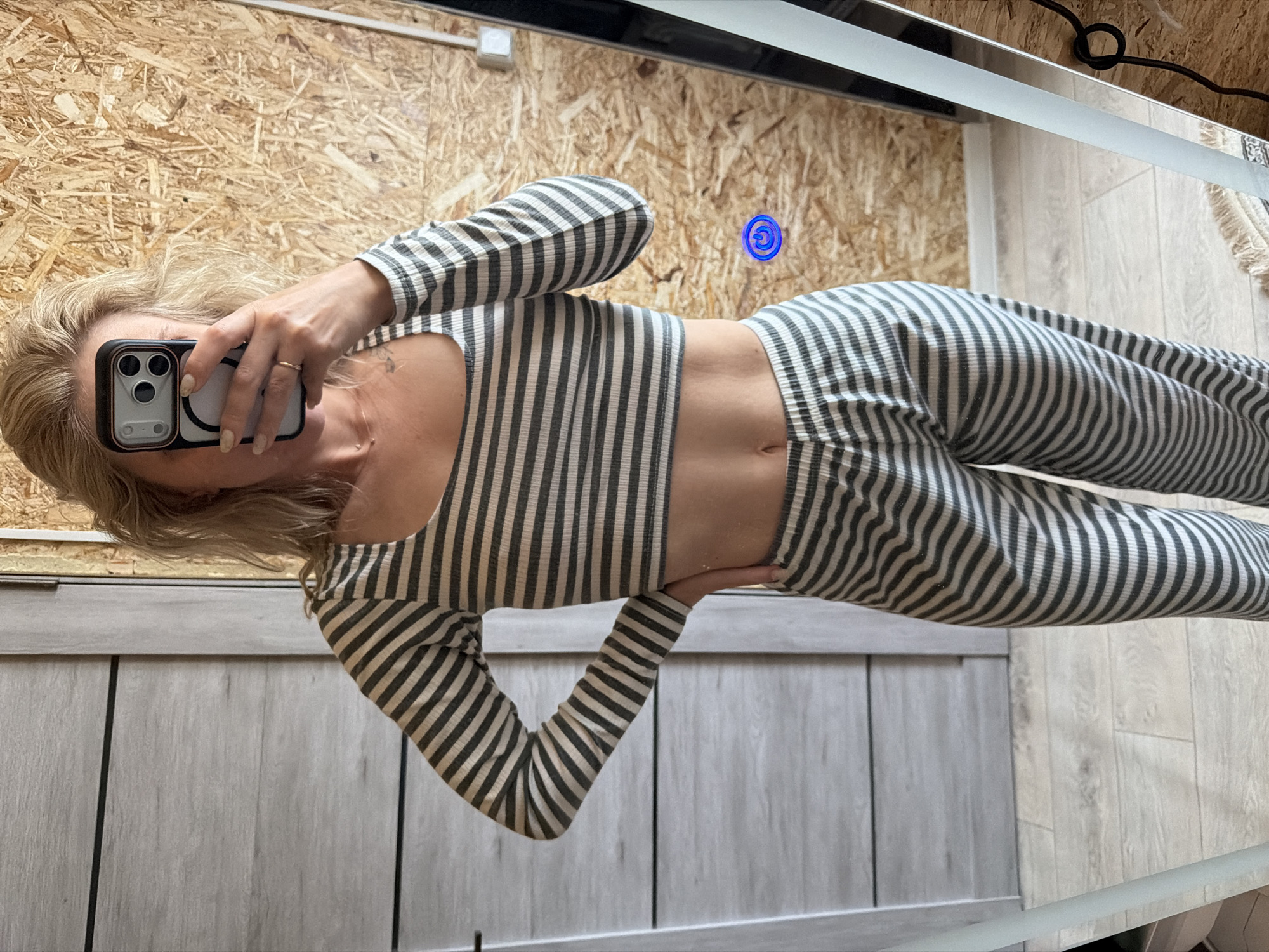 A woman is taking a selfie in front of a mirror, wearing a striped long-sleeved crop top and matching pants. The background shows a wall with a plywood texture and another wall with gray vertical panels. The woman is holding a smartphone in her right hand, partially covering her face. A blue circular object is visible on the plywood wall, and electrical cords are seen near the top of the mirror. It's not possible to determine the location as Chaikovsky, Russia, from the image.