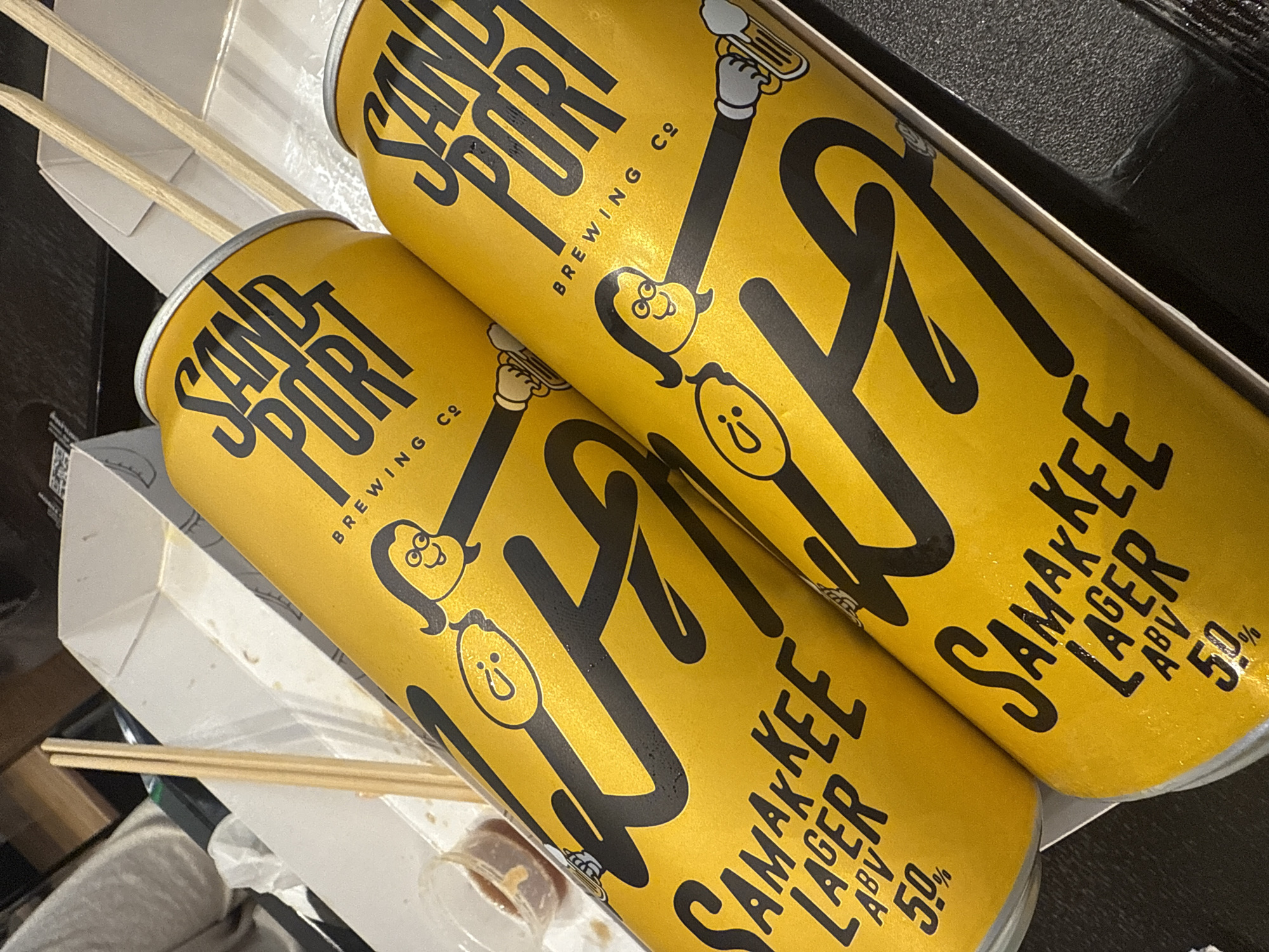 The image shows two cans of Sand Port Brewing Co. Samakkee Lager, each with an ABV of 5.0%. They are resting in a cardboard box, possibly a takeout container, alongside wooden chopsticks. A small condiment container is also visible in the box.