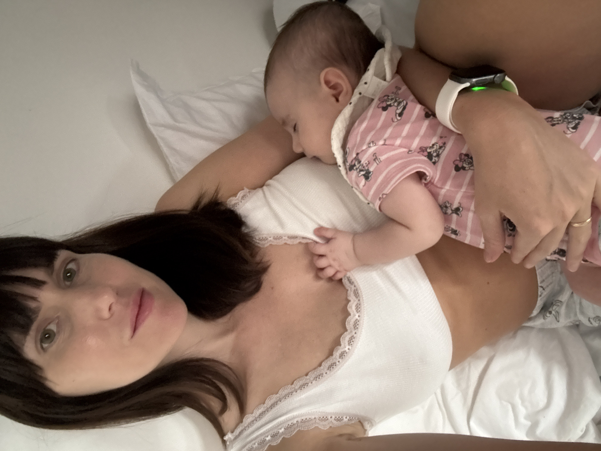 An adult with dark hair and bangs, wearing a white lace-trimmed bralette and patterned pajama bottoms, is holding a sleeping infant. The infant is wearing a pink striped onesie with cartoon characters and a white bib. The adult's left hand is on the infant's chest, and a ring is visible on the adult's ring finger. The adult is also wearing a white smartwatch on their left wrist. They are lying down on a white surface, likely a bed, with a white pillow behind them. The background is a plain, light-colored wall. The image appears to be a personal photograph taken from a slightly elevated angle. The location is not discernible from the image.
