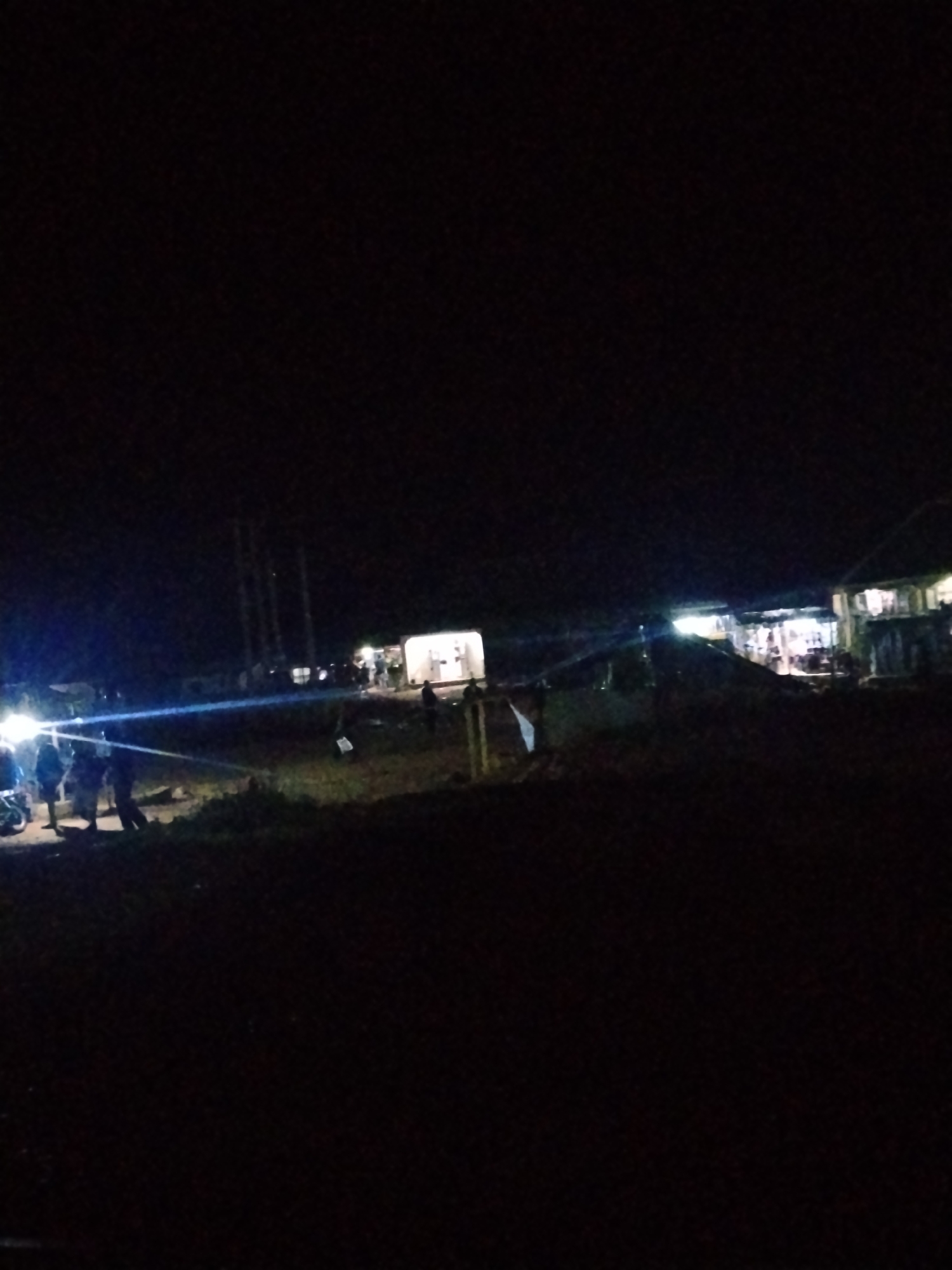The image depicts a nighttime scene in Durumi, Nigeria. The foreground is dark, with a hint of foliage. In the mid-ground, several individuals are gathered near a motorcycle with its headlights on, casting a bright beam. To the right, there are structures that appear to be stalls or small buildings with lights illuminating them, suggesting a market or gathering place. The background shows more structures and the dark night sky, with some tall, thin objects that could be utility poles. The overall impression is of a community event or an area that is active at night.