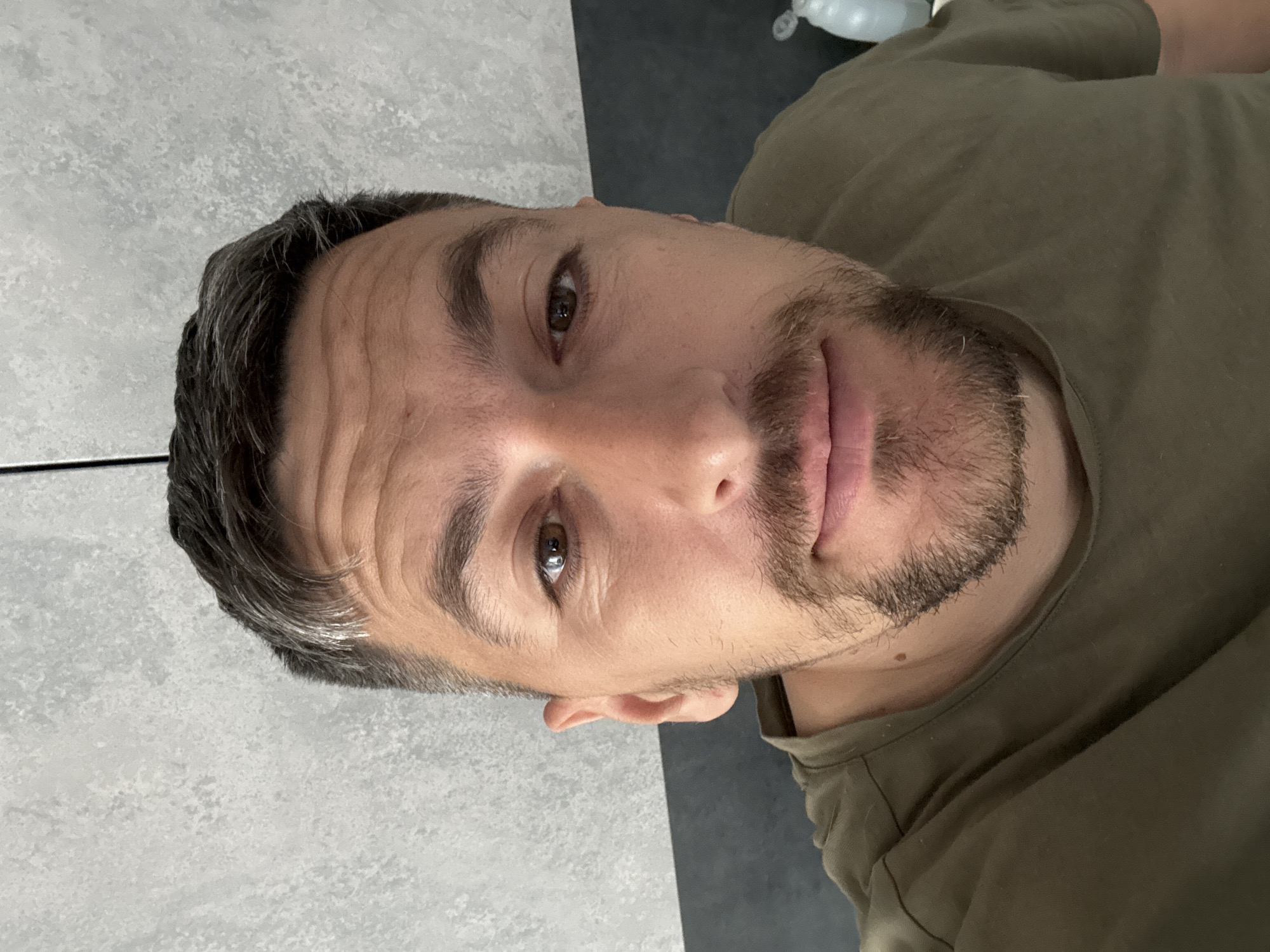 A close-up, slightly angled headshot of a man with fair skin, brown eyes, and dark hair with some graying at the temples. He has a neatly trimmed beard. The man is wearing a green t-shirt. The background is a mottled gray wall. The location is not evident from the visual information provided.