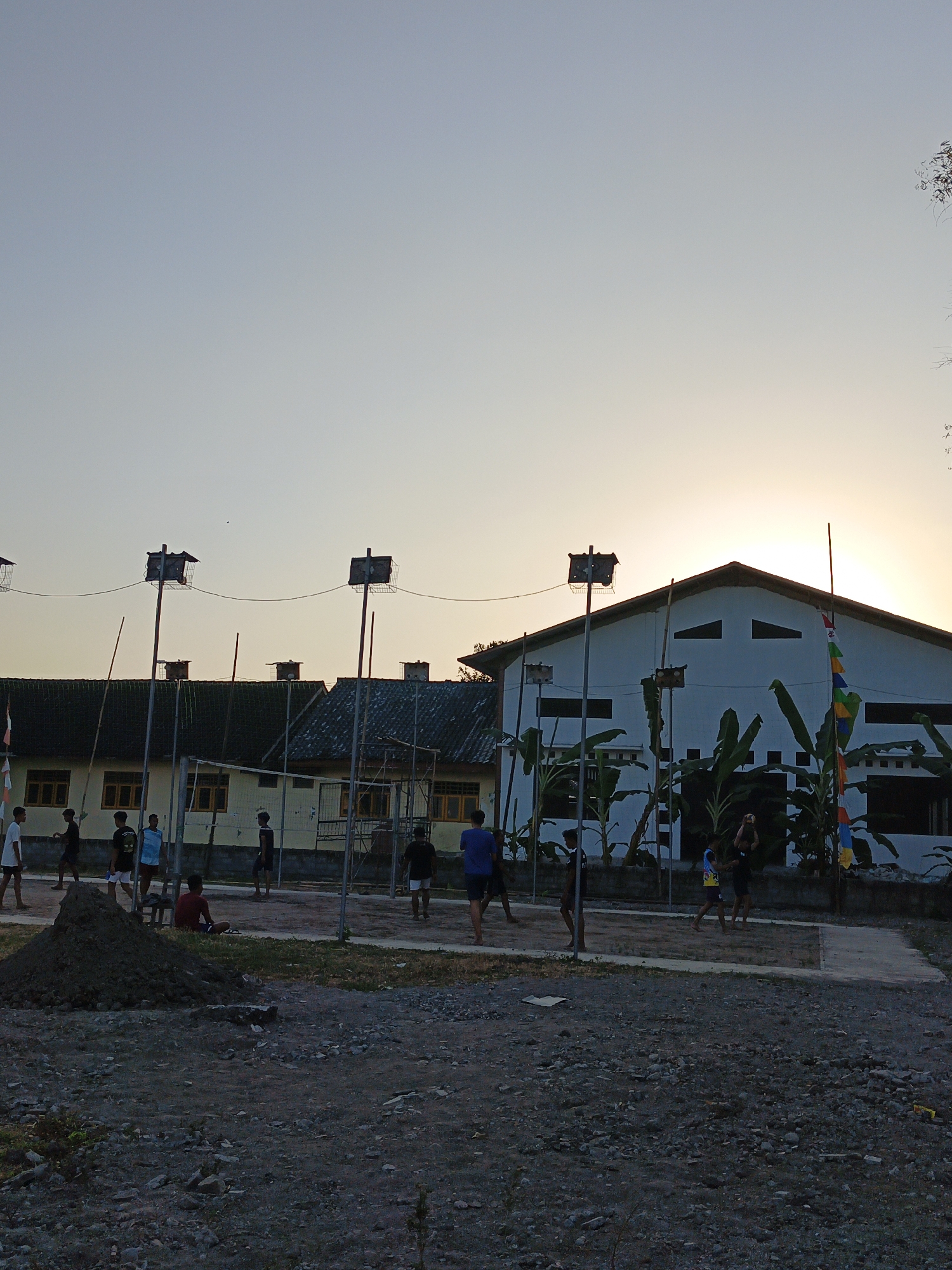 The image depicts an outdoor scene at dusk, likely in Trucuk, Indonesia, with a group of young males engaged in a game of volleyball. The setting includes a volleyball court with a net, surrounded by dirt and gravel. In the background, there are several buildings, one of which is a large, white structure with a gabled roof, and another appears to be a lower, yellow-building complex. Elevated structures with cages are visible on poles, possibly for birds. Banana trees and some flags are also present. The lighting suggests either sunrise or sunset, with a hazy, warm glow. The people are casually dressed, and their interactions are centered around the volleyball game.