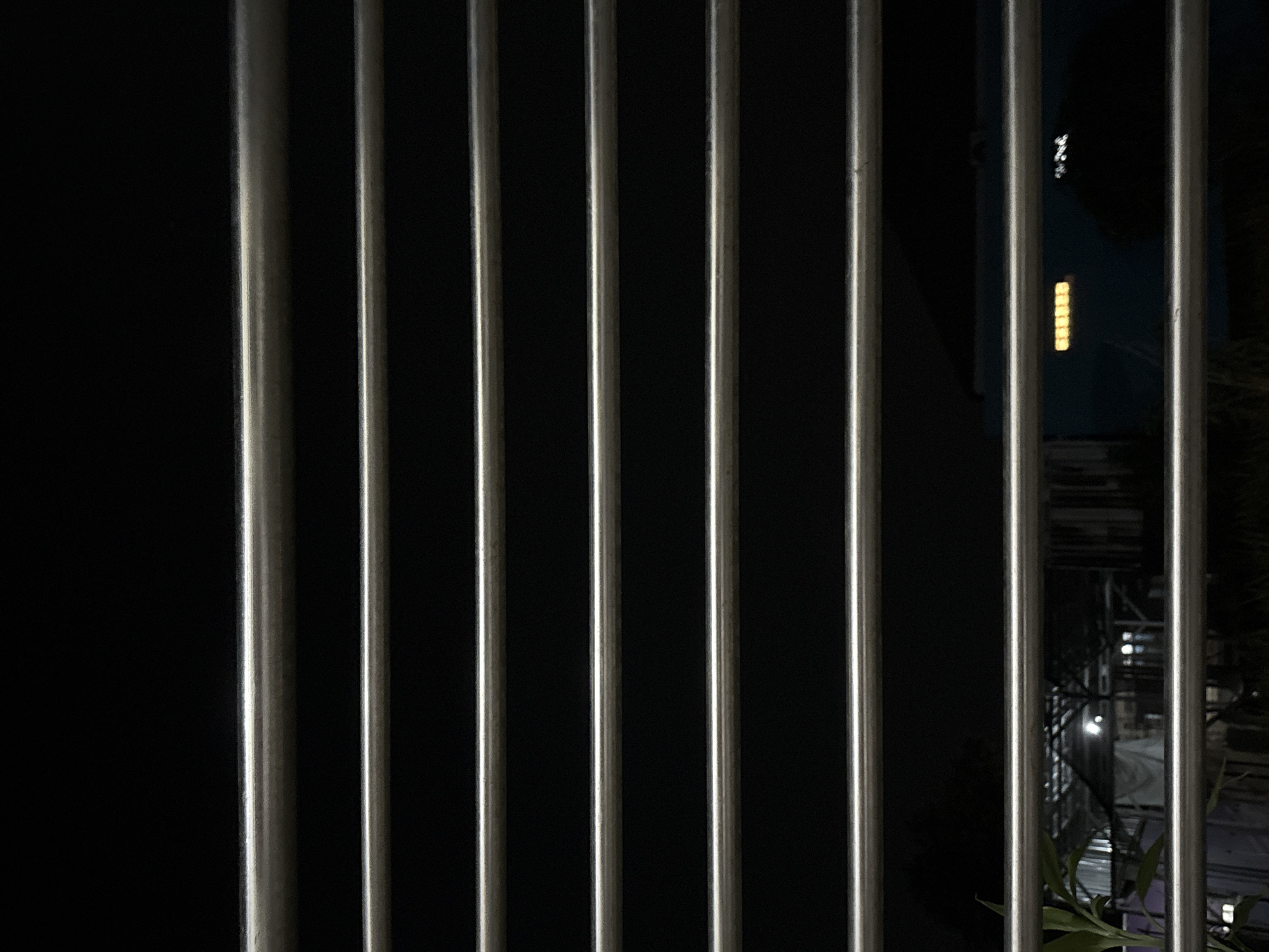 The image is a close-up shot, taken at night, of horizontal metal bars against a dark background. Through the gaps between the bars, a dimly lit outdoor scene is visible. In the background, there are structures resembling a building or a shelter with a corrugated metal roof. Lights are visible within some of these structures, and a row of yellow lights, possibly from a sign or window, is illuminated. There are also some plants or foliage in the foreground, to the left of the image, partially obscuring the view. The overall impression is of looking out from behind a barrier at a nighttime scene in what appears to be a residential or industrial area. The image is taken in Gempol, Indonesia.