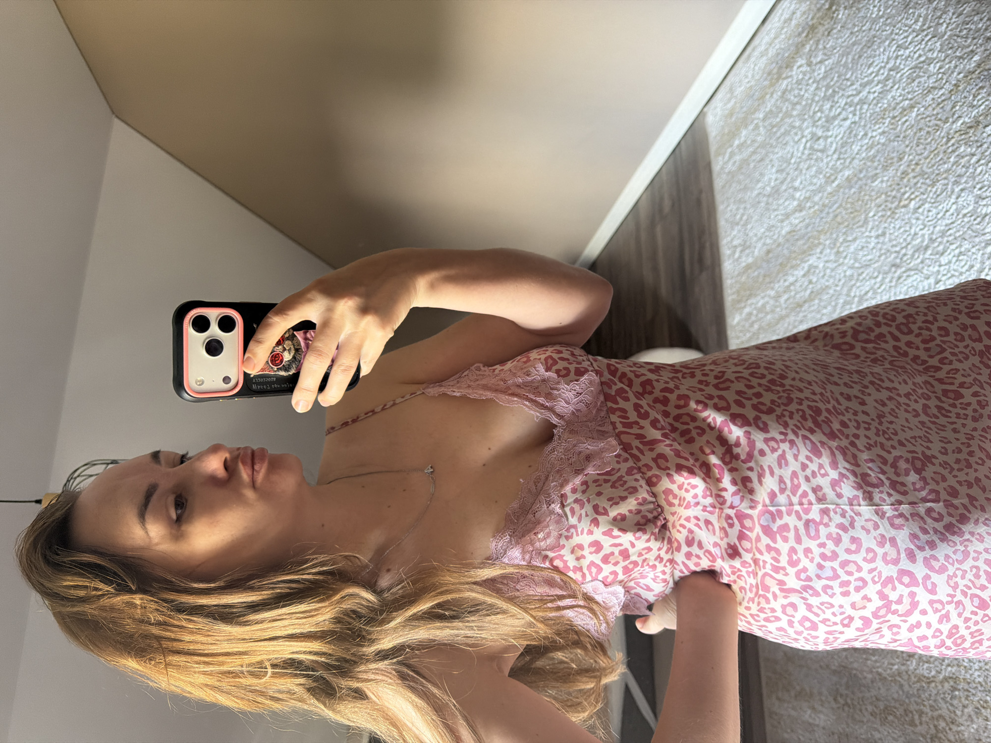 In Malinovka, Russia, a woman with long, blonde hair stands in what appears to be a bedroom. She is taking a selfie in a mirror, holding a black and pink phone with a decorated case. She is wearing a pink and white leopard print slip dress with lace trim. Her left hand is on her hip. The room has light brown walls, a white baseboard, and a wooden floor covered partly by a cream-colored textured rug. Natural light streams in from the right, casting shadows.