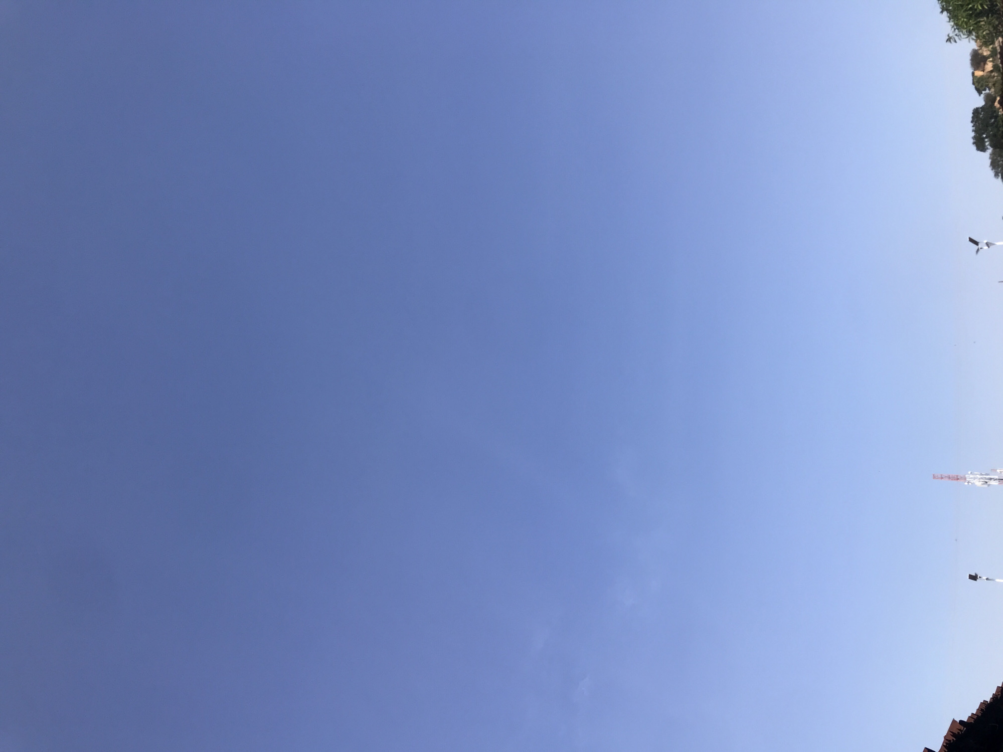 The image shows a clear blue sky with a few wisps of clouds. In the lower portion of the image, there are some rooftop structures and a tall telecommunications tower. There are also some trees visible on the right side of the image. The scene appears to be an outdoor view from a city or town.