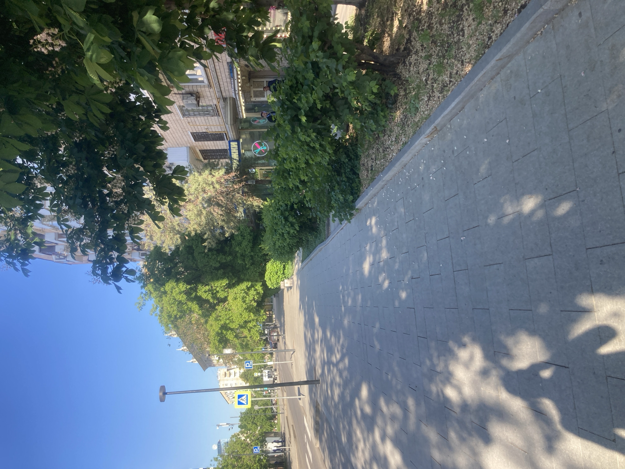 The image depicts a sunny street scene in Moscow, Russia. The perspective is from the sidewalk, looking down the street lined with buildings and trees. The sidewalk is paved with gray rectangular stones. The street itself is visible with road markings, parking signs, and streetlights. On the left, buildings with multiple floors are partially obscured by trees. A building closer to the viewer is characterized by its light-colored brick facade, air conditioning units, and signage. Along the right side of the sidewalk, a strip of greenery separates it from the buildings. The sky is a clear blue. Tree shadows dapple the sidewalk.