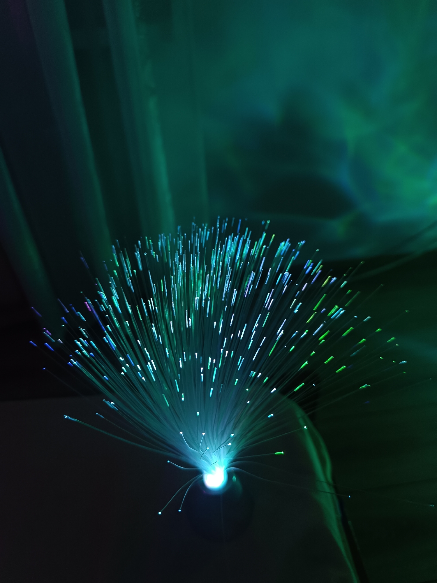A close-up, low-angle, indoor shot depicts a fiber optic lamp emitting green and blue light in a dark environment. The lamp is positioned in the lower center of the frame, with numerous illuminated fibers radiating outward and upward. The background is dark with blurred green vertical streaks possibly from curtains or other light sources.
There is no evidence to support the location as Cherkovitsa, Bulgaria.