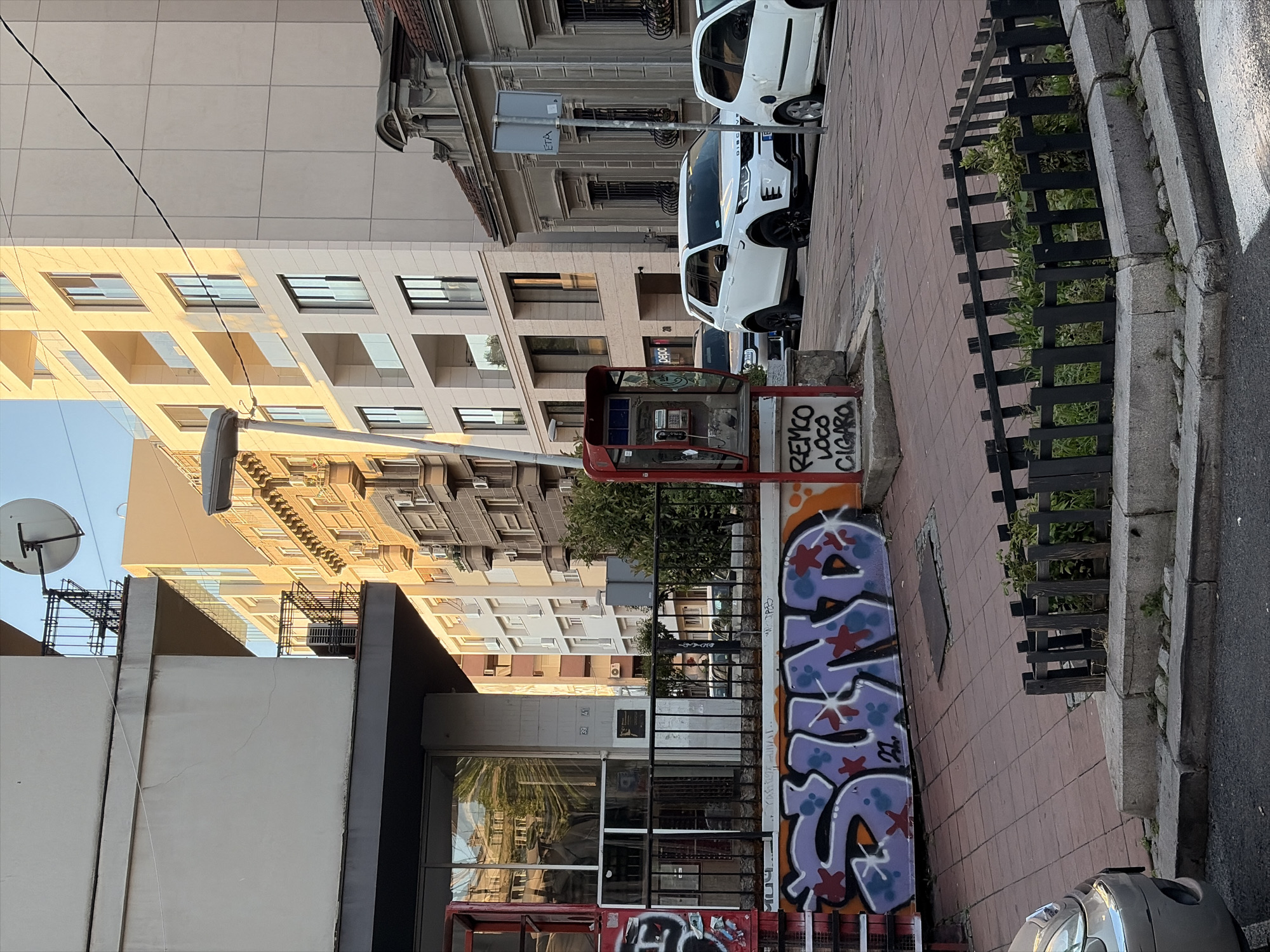 The image depicts a street scene in Belgrade, Serbia, likely in an urban residential area, possibly near a commercial district. The foreground features a red public telephone booth with graffiti on its base that reads "REMCO LOCO CIGARO". Next to the telephone booth, a colorful graffiti tag with the word "SHNO" is spray-painted on a wall.
To the left, a building with a dark gray base and a lighter upper section is visible, featuring windows and a metal fence. Beyond this building, a modern apartment building with beige facades and numerous balconies with glass railings dominates the background. Sunlight illuminates the facade of this building, casting shadows.
To the right of the telephone booth, two white cars are parked on the street. The one closer to the telephone booth is a white SUV, and further down the street, another white car is partially visible. Behind the cars, an older building with classic architectural details, including arched windows and decorative molding, stands adjacent to the modern apartment building.
The street is paved with red brick tiles, and a small, fenced-off green space with plants is situated on the sidewalk. The sky is clear and blue, indicating fair weather. The overall impression is one of a typical urban environment with a mix of old and new architecture, street art, and everyday city life.