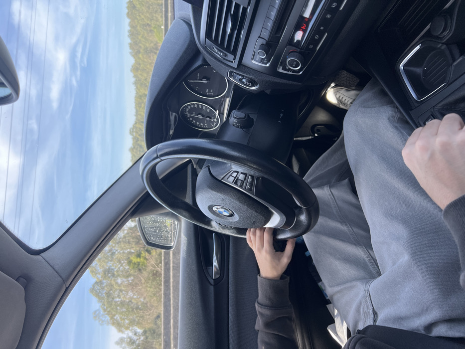 The scene depicts the interior of a BMW vehicle in motion, located in Águas Santas, Portugal. The perspective is from the front passenger seat, looking towards the driver and the road ahead.

The driver, partially visible, wears a dark grey long-sleeved top and light grey denim jeans. Their left hand grips the black multi-function steering wheel. Their right hand rests on the automatic gear shifter in the center console. The driver's right foot, encased in a light-colored sneaker, is visible near the accelerator pedal area.

The car's dashboard features two main dials: a speedometer indicating approximately 100 km/h and a tachometer showing around 2000 RPM. Below the right-side air vent, a climate control panel displays the number "2" in red, likely indicating temperature. The driver's side door panel is dark grey.

Through the windshield, a blue sky with scattered thin clouds is visible. A dense line of light green and brown trees forms the distant background, consistent with a roadside environment. A grey concrete barrier runs alongside the road on the driver's side. Faint power lines stretch across the upper part of the sky. The driver's side mirror exhibits water droplets or marks on its surface.
