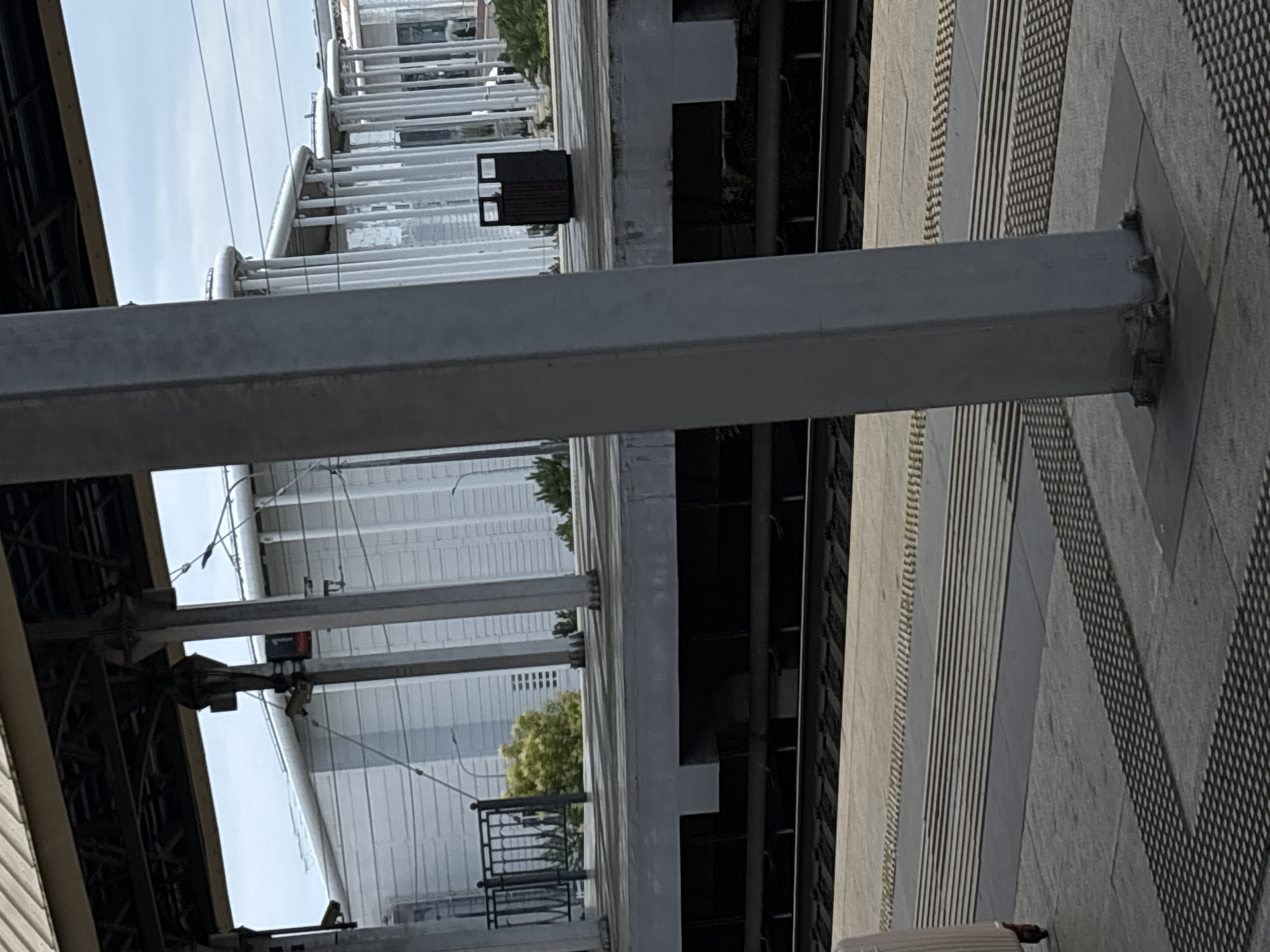 The image shows a train station platform in Sirius, Russia. The platform has a gray surface with tactile paving for visually impaired pedestrians. There are train tracks visible in the foreground. Behind the platform, there are several large white buildings with a ribbed facade and cylindrical structures on top. A dark gray trash bin is located on the platform. A large gray pillar obstructs the view of the station and the buildings in the background. The sky is overcast and appears to be daytime.