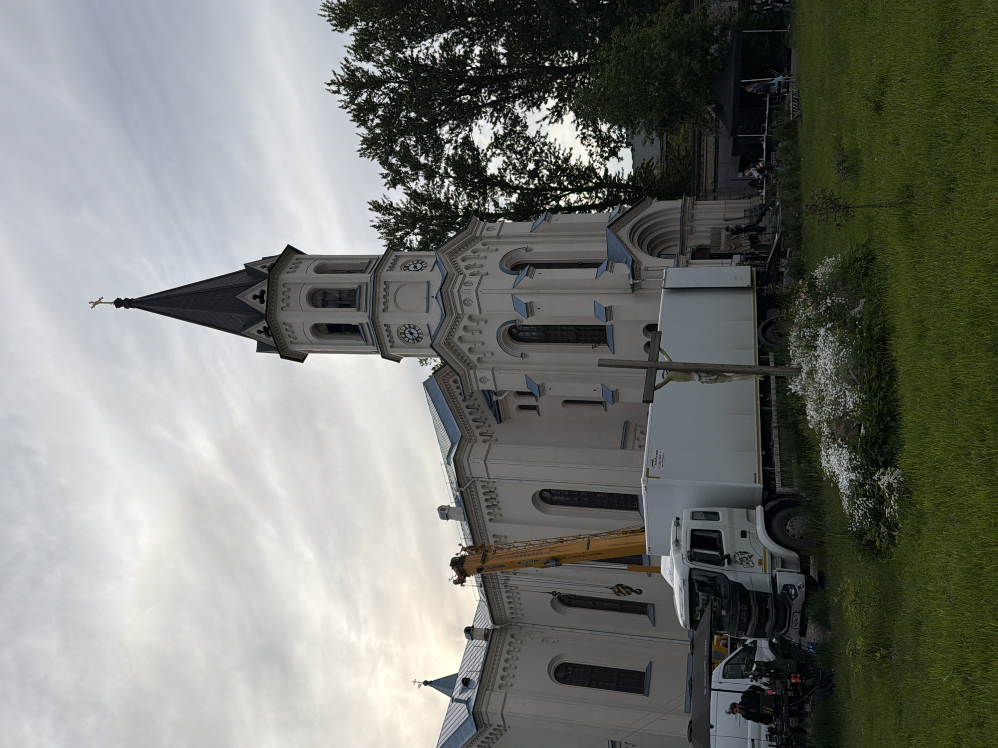 The image displays an outdoor scene dominated by a large, ornate church building under a cloudy sky. The church features light-colored walls, possibly white or cream, and a tall, dark-spire tower topped with a golden cross. The tower is adorned with multiple round clock faces and intricate architectural details, including arched windows and decorative cornices.
In the foreground and mid-ground, a significant amount of equipment and several vehicles are present, suggesting a production setup or event. A prominent yellow mobile crane truck is positioned with its boom extended, alongside multiple white box trucks. One of these trucks has Russian text on its side which reads, "Аренда автоподъемника 20-45 метров. От 1000 руб/час" (Car lift rental 20-45 meters. From 1000 rub/hour). The cab of another truck clearly displays the "JAC" logo. Various other professional equipment, including what appears to be camera gear and lighting, is scattered around the area.
To the right of the church, a well-maintained grassy lawn extends, featuring small flowering bushes with white blossoms and a slender young tree. Further in the background, a dark stage-like structure is visible, with several people standing on or near it. More individuals are seen around the vehicles and equipment, engaged in various activities, indicating an ongoing setup or event. Large, leafy trees frame the scene against the sky.
The weather appears to be overcast or partly cloudy, characterized by soft, diffused daylight and streaks of pale clouds. The overall impression is that of a bustling outdoor production or event taking place against the backdrop of a historic religious building in Saint Petersburg, Russia.