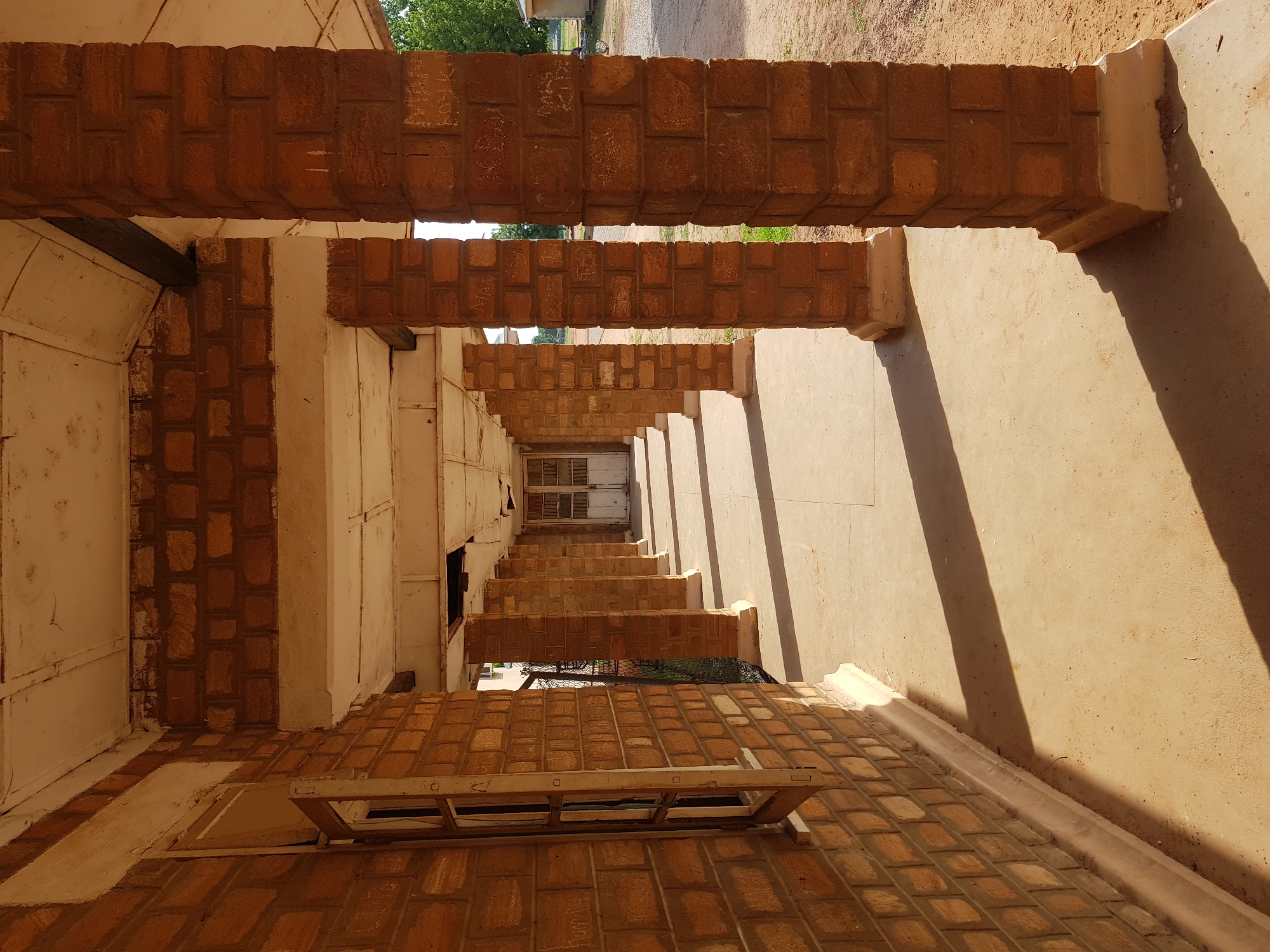 This image depicts a long, covered walkway supported by a series of brick pillars. The walkway appears to be part of an older building, possibly a school or administrative facility, located in Jimeta, Nigeria. The brickwork of the building and the pillars is a warm, earthy tone, with some visible signs of age and wear. The roof of the walkway is white and shows some damage, with sections of ceiling missing. Sunlight streams in from the right, casting long shadows across the concrete floor. The walkway extends into the distance, leading towards a set of closed double doors at the far end. To the left of the walkway, a brick wall with a single, narrow window is visible. Outside the walkway, to the right, a dirt ground and some green trees can be seen, suggesting an outdoor environment. There are no people visible in the image. The overall impression is one of a functional but aged structure in a warm, sunny climate.