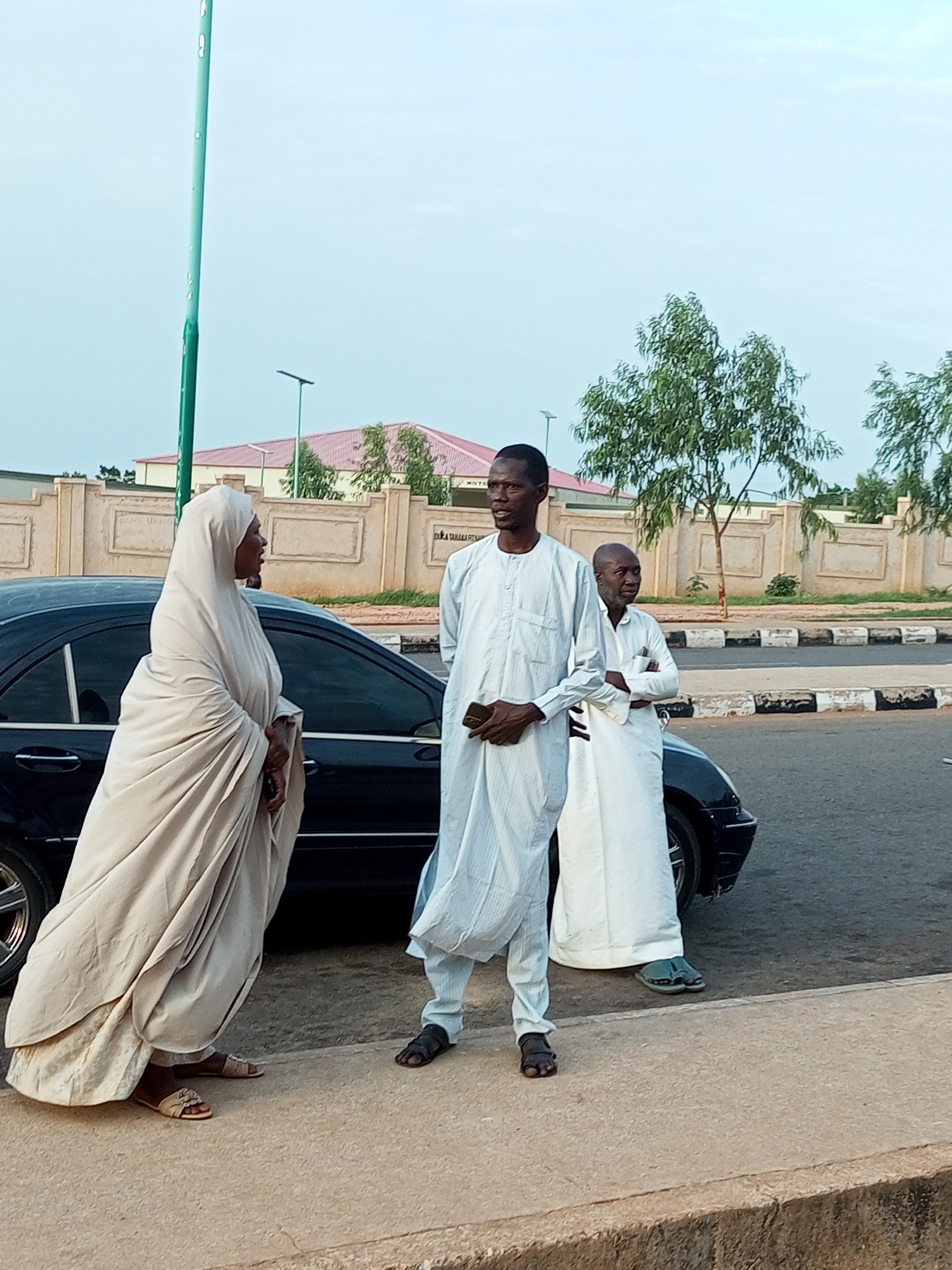 Three people stand next to a black Mercedes-Benz sedan on the side of a road in Gombe, Nigeria. On the left, a woman in a light-colored hijab and dress stands facing away from the camera, her right hand resting on her hip. To her right, a man in a light-colored tunic and pants holds a smartphone in his left hand and faces the woman. Another man, also in a light-colored tunic and pants, stands behind the first man, facing the camera and with his arms crossed. The car is parked on the asphalt road, with a concrete sidewalk in the foreground. In the background, a beige wall with decorative fencing lines the road, and behind it are buildings with red roofs and trees. The sky is overcast.