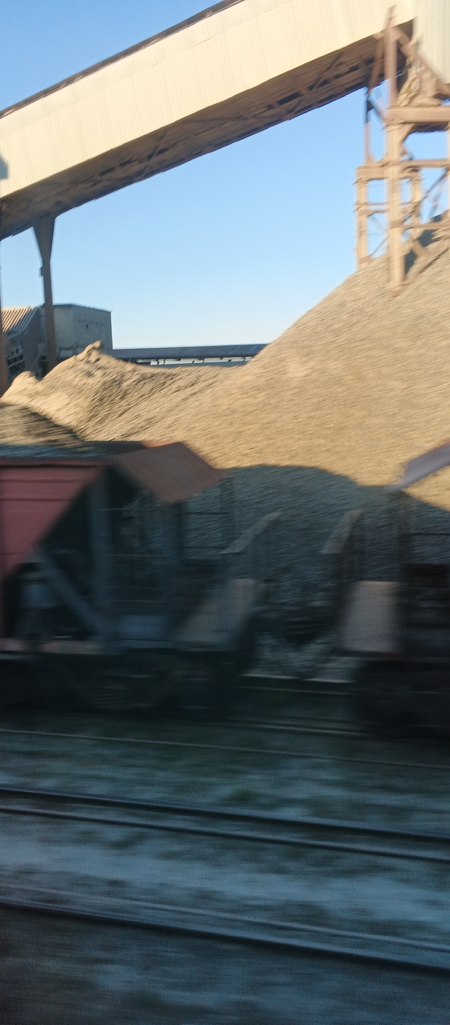 In Khlystunivka, Ukraine, this image captures an industrial scene, likely a quarry or construction site, under a clear blue sky. A large, beige conveyor belt system extends across the upper portion of the frame, supported by a weathered metal structure on the right. Below the conveyor belt, massive piles of light brown gravel or crushed stone dominate the midground. The gravel mountains are immense, suggesting a significant operation. In the foreground, a train of freight cars is visible. The cars are dark, some with red or brown elements. The motion blur indicates the photo was taken from a moving vehicle, likely the train itself. The tracks in the very bottom of the image are also blurred, further emphasizing movement. The scene is devoid of people.