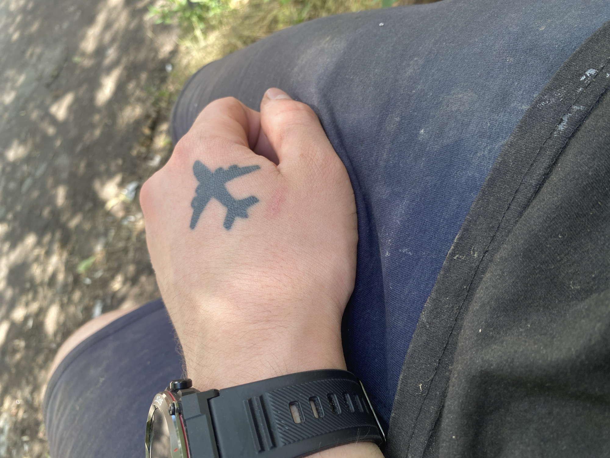 The media file shows a close-up view of a person's hand and arm, featuring a black airplane tattoo on the back of their hand. The person is wearing a black watch with a ribbed strap and a red accent on the bezel. They are also wearing dark blue shorts or pants and a black garment, possibly a shirt or jacket, with a ribbed edge. The background is blurred, but it appears to be an outdoor setting with dappled sunlight filtering through leaves, suggesting a daytime scene. The lighting indicates it is likely sunny. The focus is on the hand and the tattoo, highlighting the details of the artwork and the watch. The overall impression is casual and personal, with the tattoo and watch being prominent personal details. There is no visible text in the image.