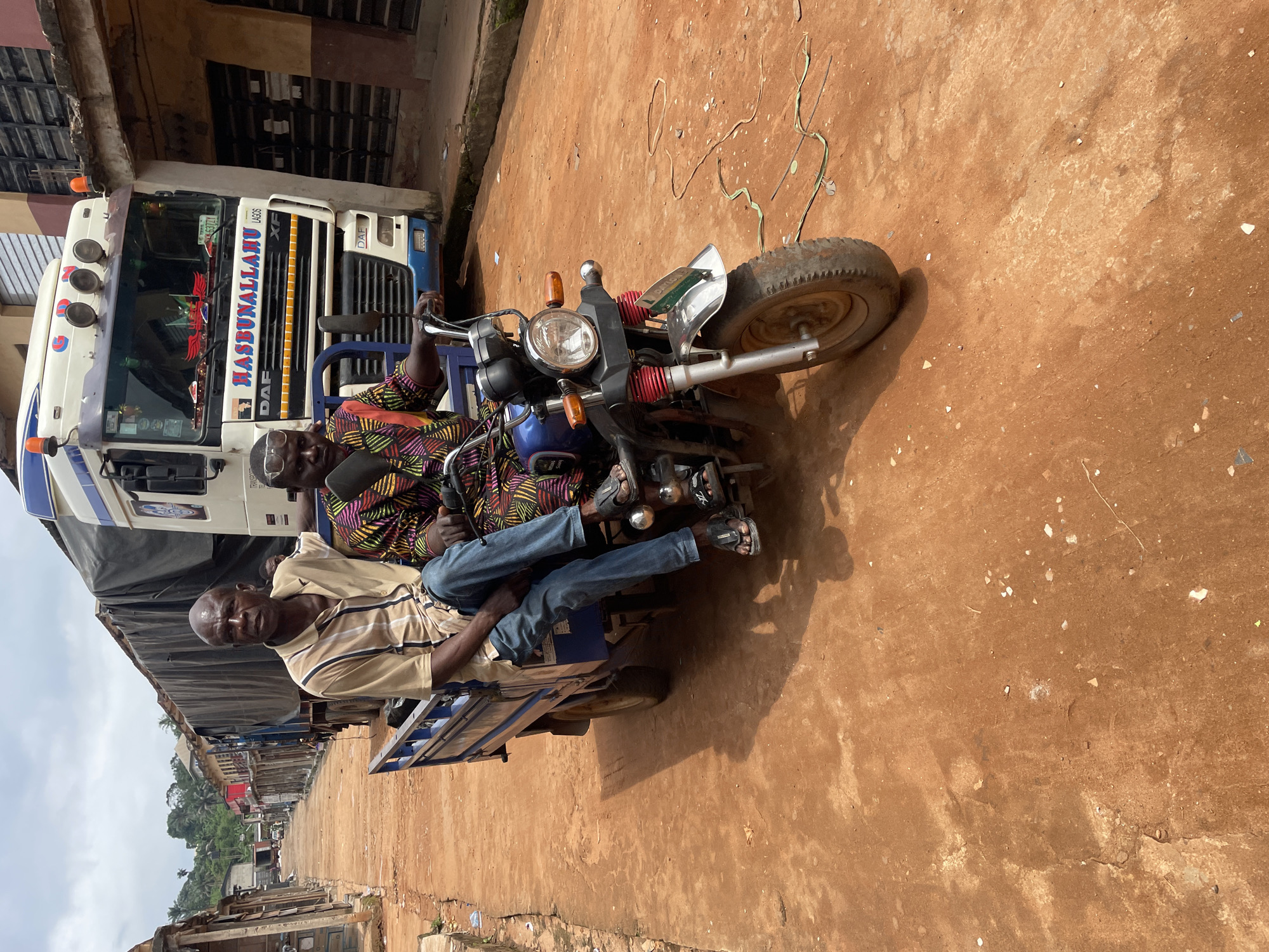 The image depicts a street scene in the city of Ogidi, Nigeria. In the foreground, two men are seated on a three-wheeled vehicle, resembling a modified motorcycle with a cargo bed. The man in the front is wearing a light-colored, striped short-sleeved shirt and blue jeans. The man behind him is dressed in a vibrantly colored, patterned shirt. Both men are wearing sandals. The vehicle has a blue cargo bed and a silver fender. Behind the men and their vehicle, a large truck with a tarp-covered cargo is parked. The truck appears to be a DAF model, with "HASBUNALLAHU LAGOS" written on its side. The street is unpaved and appears to be made of reddish-brown earth. Buildings line the sides of the street, some with weathered facades and corrugated iron roofs. In the background, trees and lush vegetation are visible, suggesting the area is not entirely urban. The sky is overcast with scattered clouds. The overall impression is of a busy, functional street in a Nigerian city.