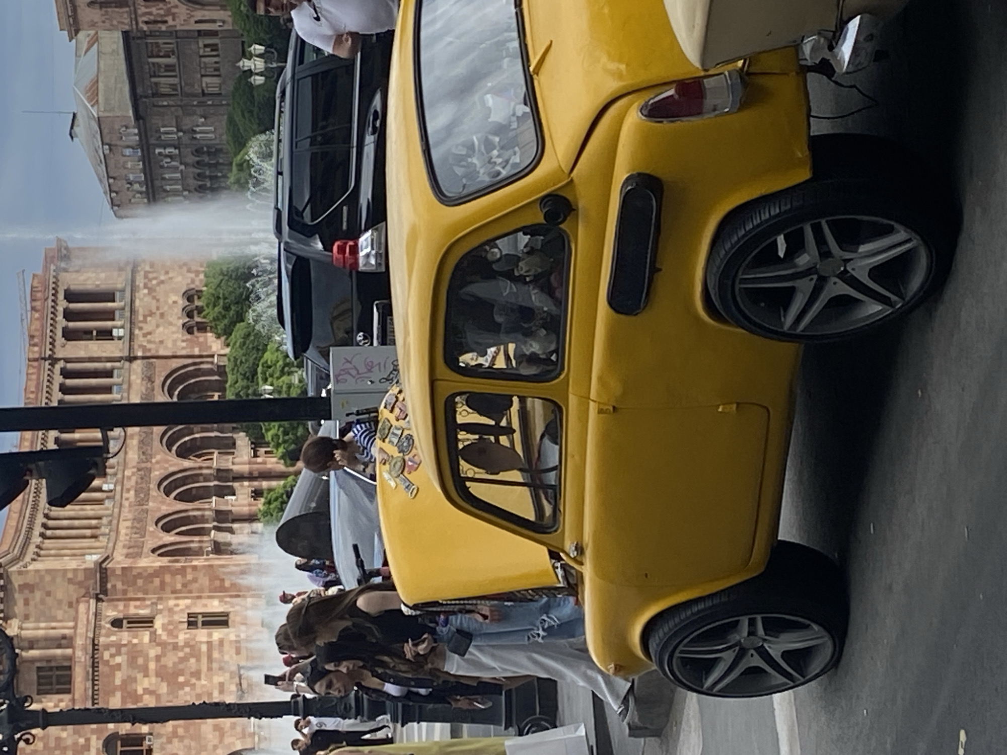 The image captures a bustling daytime scene in Yerevan, Armenia, centered around a distinctive, bright yellow, custom-modified vintage-style microcar. The car, possibly a Fiat 500 or similar model, is prominently featured on the right side of the frame, showcasing modern, five-spoke alloy wheels that contrast with its classic body. Its hood is decorated with various stickers, some appearing to be currency or travel-themed.
Behind the yellow car, a dark-colored SUV is parked, with some illegible graffiti-like text, including "G4L", visible on its rear. The background is dominated by grand, reddish-brown stone buildings with numerous arched windows and balconies, characteristic of Yerevan's architecture, particularly around Republic Square. An active fountain with multiple water jets rises between these buildings, creating a misty effect.
Numerous people are visible throughout the scene, particularly around the yellow car and in the mid-ground. They appear to be casually observing, walking, and some are seemingly taking photos, suggesting the car is an attraction at a public gathering or street event. One person in a striped top is seen near the front of the yellow car, and others are scattered throughout the square.
The setting is a public square or street, under what appears to be diffused daylight, indicating fair weather. The overall mood is lively and curious, typical of a public event or a popular urban gathering place. Visible text is limited to the partially decipherable "G4L" on the SUV's rear and indistinct patterns on the yellow car's decorative stickers.