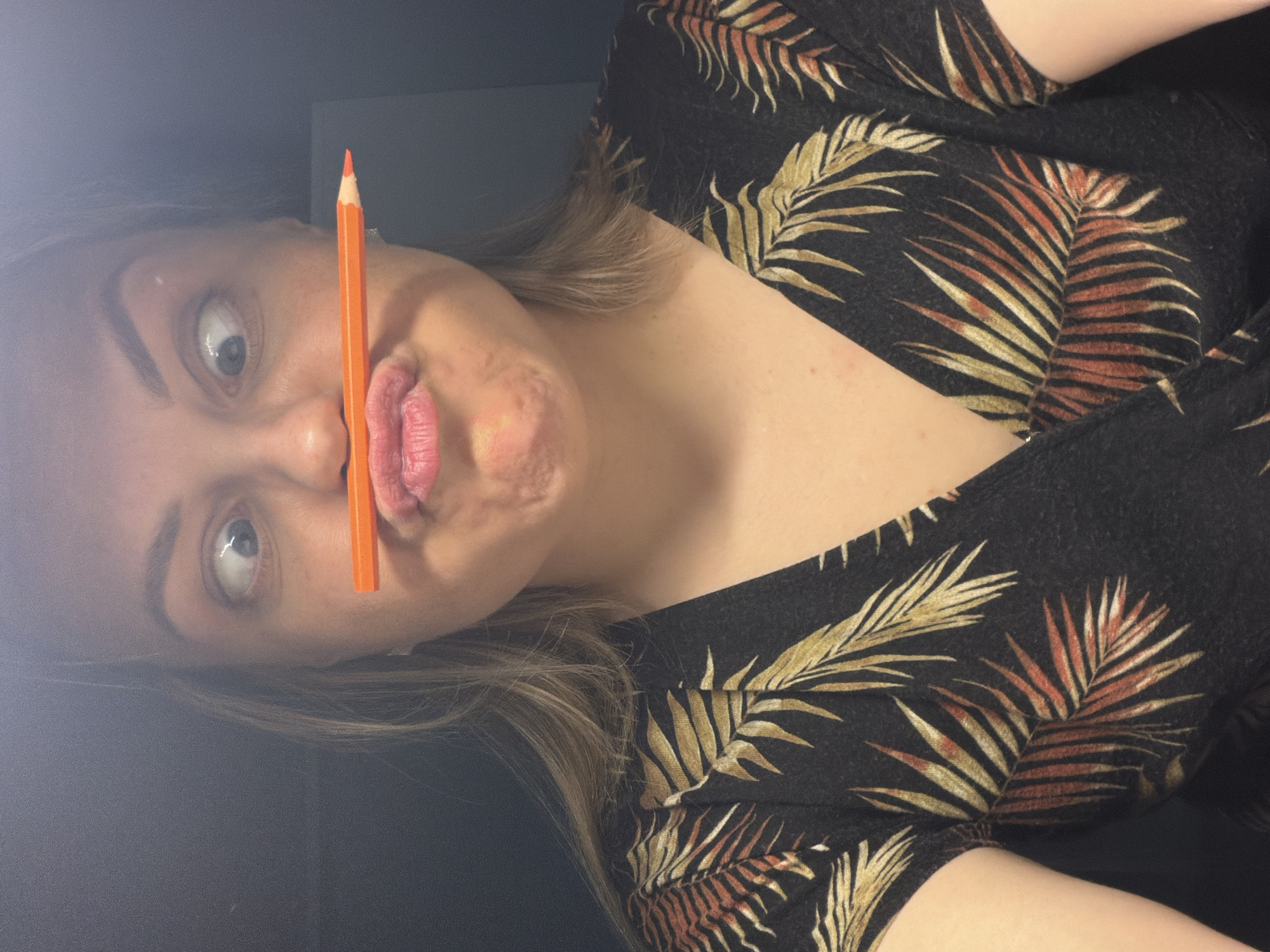 A young woman is holding an orange pencil between her nose and upper lip, making a playful face with wide eyes and pursed lips. She is wearing a dark-colored shirt with a leafy pattern. The background appears to be a dark wall. The image is rotated slightly. There is no indication of the city of Votkinsk, Russia, or any other location in the image.