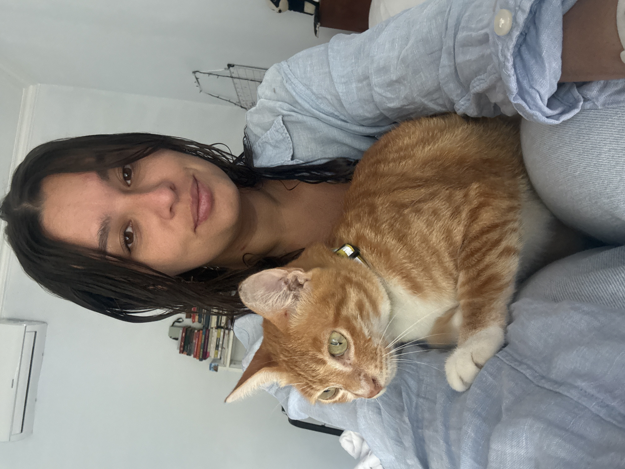 A woman with wet dark hair and brown eyes is holding an orange tabby cat with green eyes. Both the woman and the cat are lying on a light blue denim-like fabric. The woman is wearing a light blue shirt. A bookshelf with books and a wall-mounted air conditioning unit are visible in the background. The location, Pejeng, Indonesia, cannot be confirmed from the image.