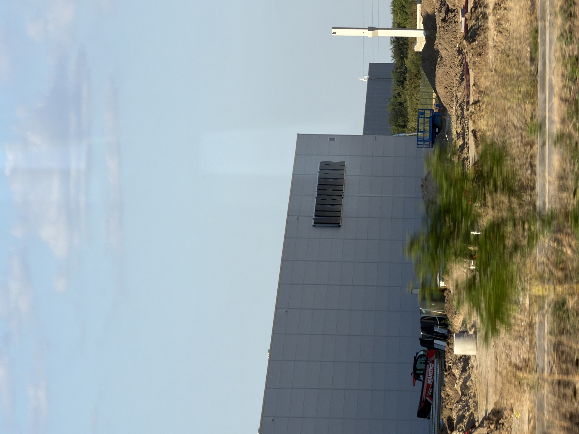 The image depicts a construction site in the city of Benkovski, Bulgaria. A large, modern warehouse-style building with "LIEBHERR" signage is prominent in the center. The building has a light gray, panelized exterior. In the foreground, construction activity is visible. A red Manitou telehandler is parked on a pile of dirt. Various construction materials, including concrete pipes and possibly scaffolding, are scattered around the site. A blue scissor lift is also present. The ground is uneven, with mounds of soil and dry vegetation. In the background, more construction appears to be underway, with another dark-colored building visible. The sky is a clear blue with a few scattered clouds. The overall impression is one of ongoing industrial development.