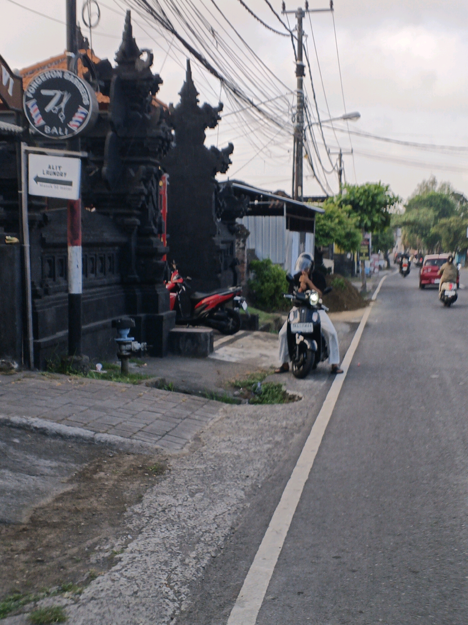 The image captures a street scene in Denpasar, Indonesia, likely during overcast conditions. In the foreground, a person wearing a helmet and white pants is seated on a black scooter, which is parked on the side of the road, facing right. The scooter's headlight is illuminated. To the left of the scooter, a dark, ornate gate structure is visible, adorned with traditional Balinese carvings. A circular sign above the gate reads "FORGERON BARBERS BALI" with stylized scissors. Below the gate, a white directional sign with an arrow pointing left indicates "ALIT LAUNDRY." A red and white striped pole stands next to the gate. A red scooter is parked near the gate.
The road itself is asphalt, marked with a white line indicating the lane boundary. Several other scooters and a red car are visible further down the road, suggesting traffic. Electrical wires are strung across the sky, supported by utility poles. Green trees and foliage are present on the right side of the road. The pavement in front of the gate appears to be made of stone tiles, transitioning to a rougher surface closer to the road. A metal pipe with blue accents is present on the sidewalk. The overall atmosphere suggests an urban or semi-urban environment in Denpasar.