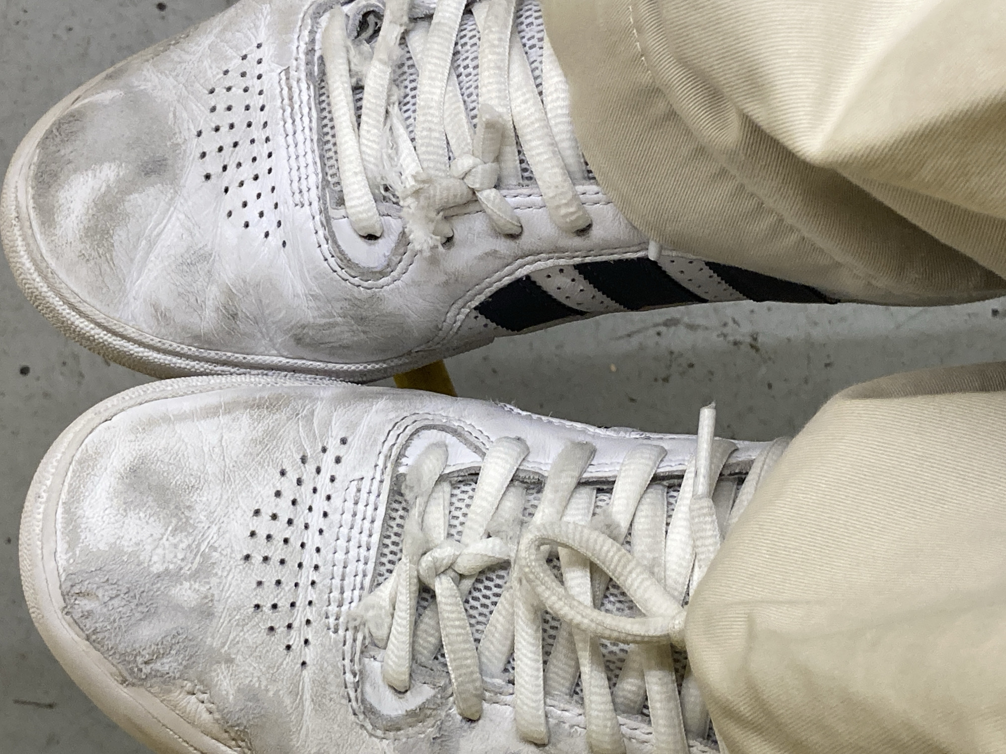 This is a close-up, overhead shot of two well-worn, white Adidas sneakers. The sneakers are made of leather and have several rows of perforations on the toe boxes for ventilation. The laces are white and appear to be slightly frayed on the right shoe. The shoes are dirty and scuffed, with visible marks and discoloration on the leather. The left shoe has a visible stitched detail along the toe. The right shoe has the iconic three black stripes of Adidas visible on the side. Both shoes are laced up and are partially obscured by beige-colored fabric, likely pants, in the foreground. The background appears to be a light gray, textured surface, possibly concrete or pavement. The lighting suggests an outdoor setting. The image does not contain any discernible elements that would place it in Barcelona, Spain.