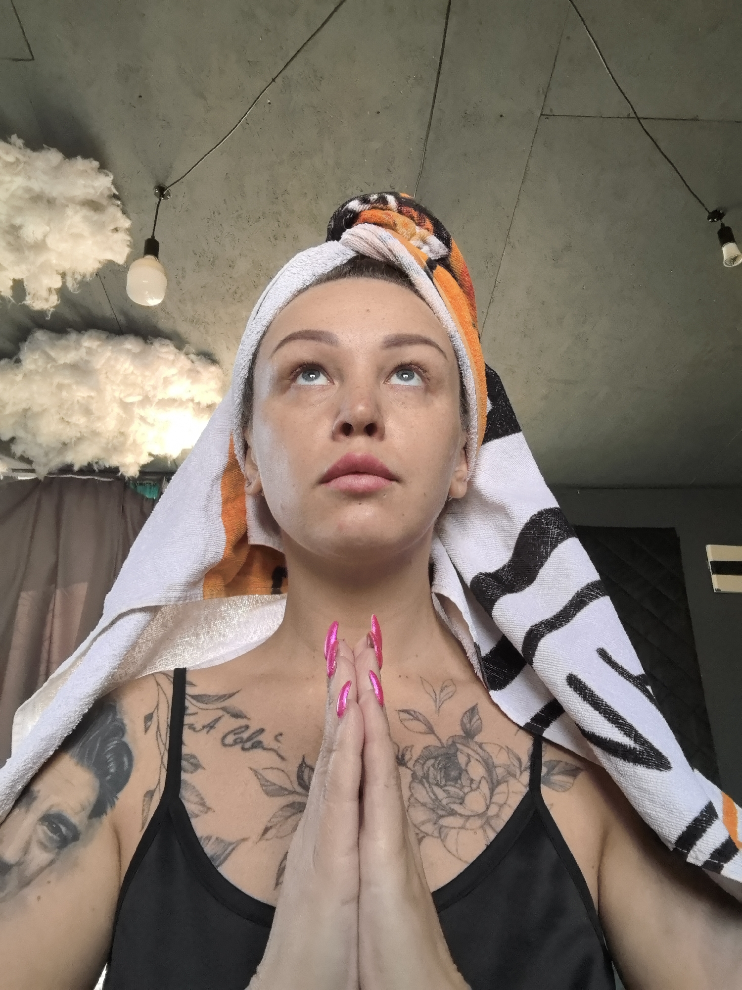 A woman is posing for the camera with a towel wrapped around her head, holding her hands together in front of her chest, as if in prayer. She is wearing a black tank top and has tattoos on her chest and arms, including a portrait of a man and a rose. The background features decorative clouds and exposed light bulbs hanging from the ceiling. It is unlikely that the image was taken in the city of Noviy, Russia, as there is nothing in the image to suggest that it was taken there.