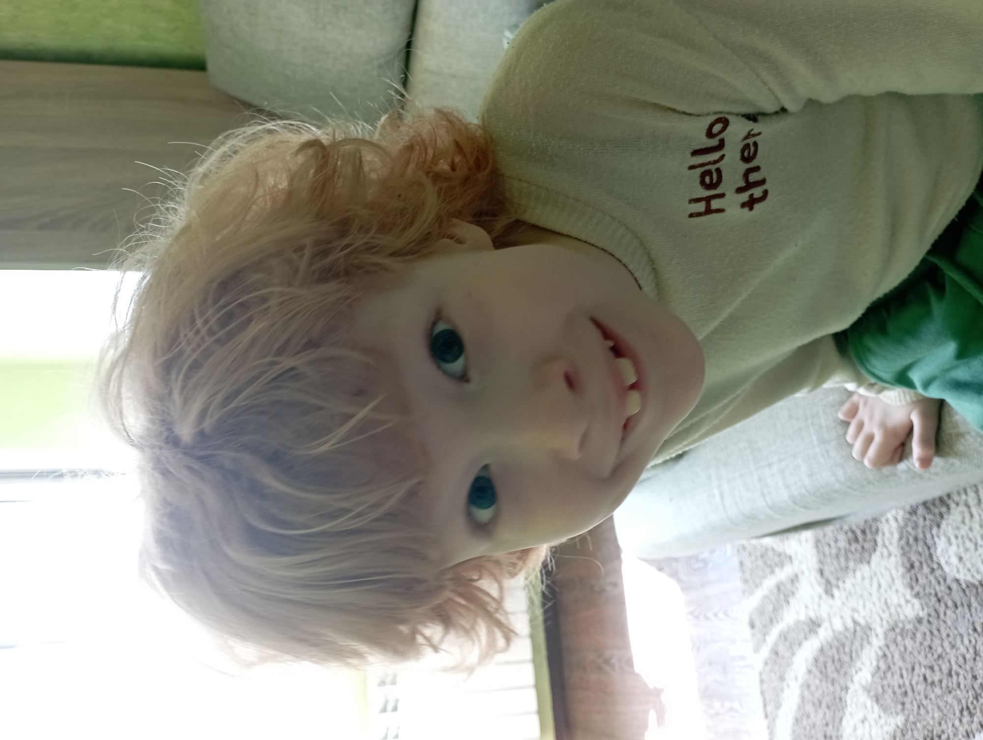 A young child with curly red hair and bright blue eyes is looking directly at the camera with a wide, toothy grin. The child is wearing a light-colored sweater with the words "Hello there" printed in dark red on the chest. The child is sitting on a light-colored couch, and a patterned rug is visible on the floor. The background is out of focus but appears to be an indoor setting with natural light coming from a window. The location is not specified in the image.