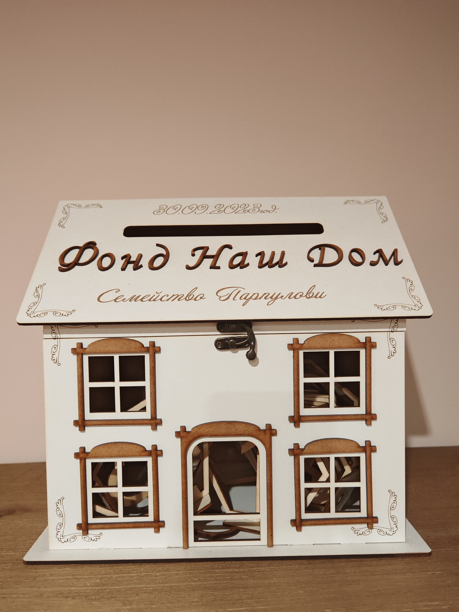 A white, wooden money box designed to resemble a house sits on a wooden surface. The house-shaped money box has a slot at the top and is decorated with brown wooden lettering and ornate scrollwork. The lettering on the front of the house reads "Фонд Наш Дом" (Fund Our Home) and "Семейство Парпулови" (Parpulo family). Below the main lettering, there is a date: "30.09.2023 год." The house has multiple windows, each framed in brown wood, and a small latch on the front door area. The background is a plain, light beige wall. The location in the city of Plovdiv, Bulgaria, is not discernible from the image. There are no people or actions depicted in the scene.