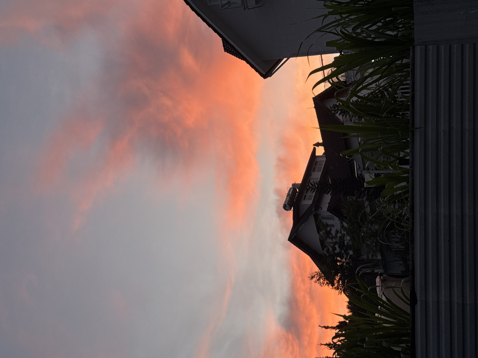 A twilight sky, painted with hues of orange and pink, forms the backdrop to a suburban scene in Lâm Viên - Đà Lạt, Vietnam. Silhouetted against the vibrant sky are several houses with tiled roofs, their structures partially obscured by lush green foliage in the foreground. The houses appear to be two or three stories tall, with visible windows and some rooftop fixtures, possibly solar water heaters. A corrugated metal fence runs along the bottom of the frame, separating the viewer from the houses and vegetation. The overall atmosphere is tranquil, characteristic of a serene evening in a residential area.