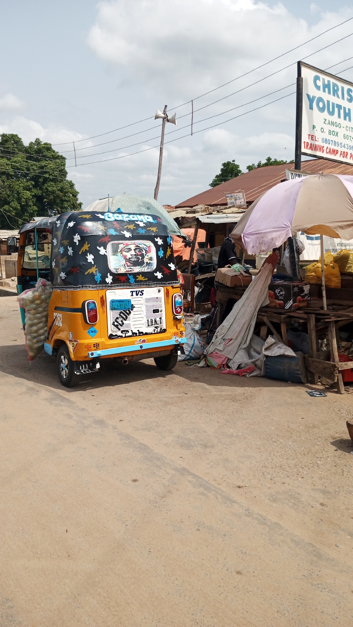 The image depicts a yellow and blue tricycle, likely a "keke" or auto-rickshaw, parked on a paved road in Zango-ndaji, Nigeria. The tricycle has a black top decorated with white and blue shapes and a portrait of a man. Bags of what appears to be produce are strapped to the side of the vehicle.
Next to the tricycle is a market stall under a pale purple umbrella. Various goods are displayed on the stall. A person is standing near the stall, possibly a vendor.
In the background is a sign for "Christ Youth," with a contact number. Utility poles and overhead wires are also visible, along with trees and buildings. The sky is overcast.