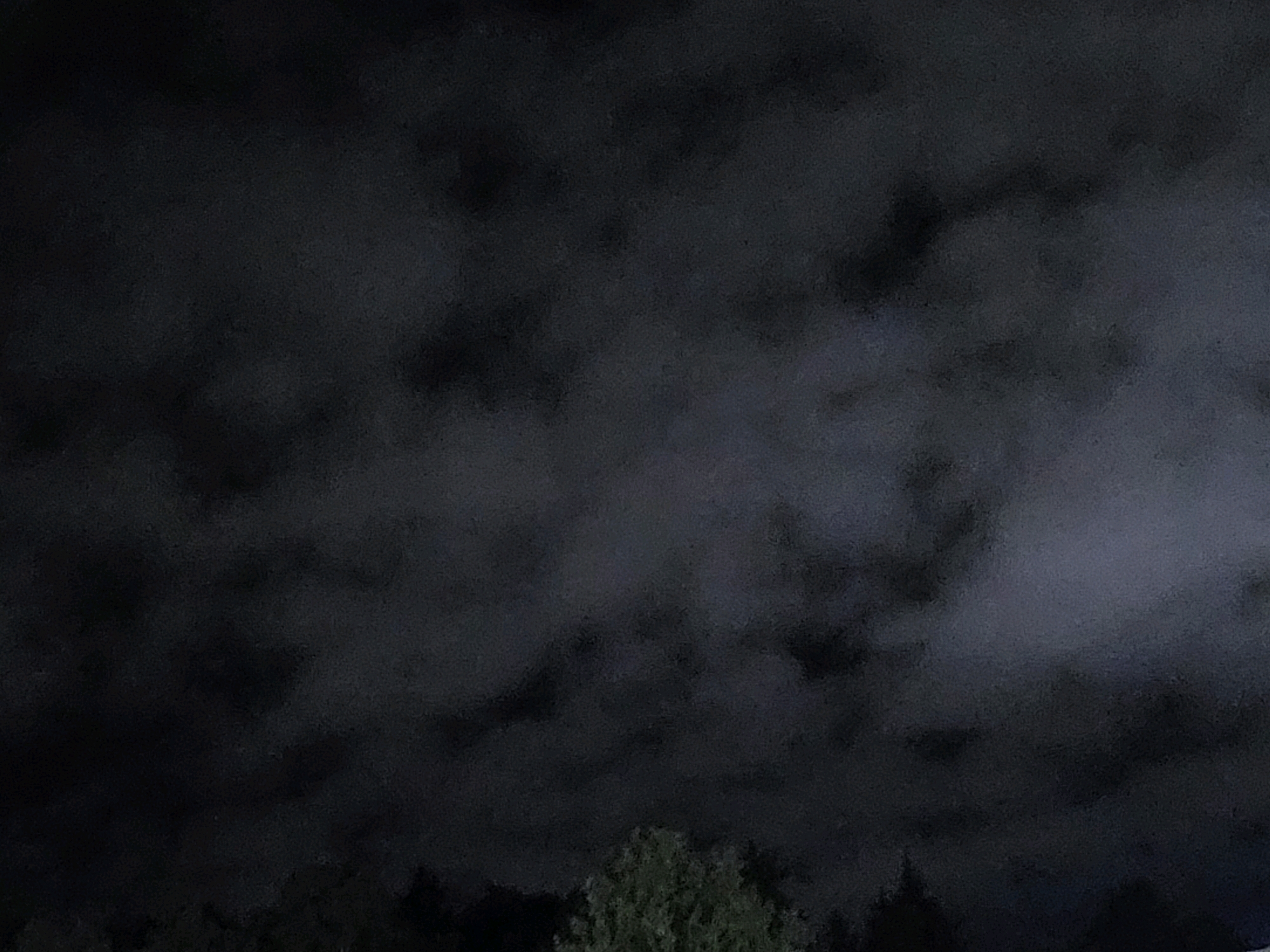 The image depicts a dark, overcast sky with visible cloud formations. The lower portion of the image shows a silhouette of trees and foliage against the sky. The scene is devoid of any people or identifiable man-made structures, suggesting an outdoor, possibly rural or natural setting. The overall impression is one of a muted and possibly stormy atmosphere. The image does not contain any visual information that would allow for the specific identification of its location within the city of Ozolnieki, Latvia.