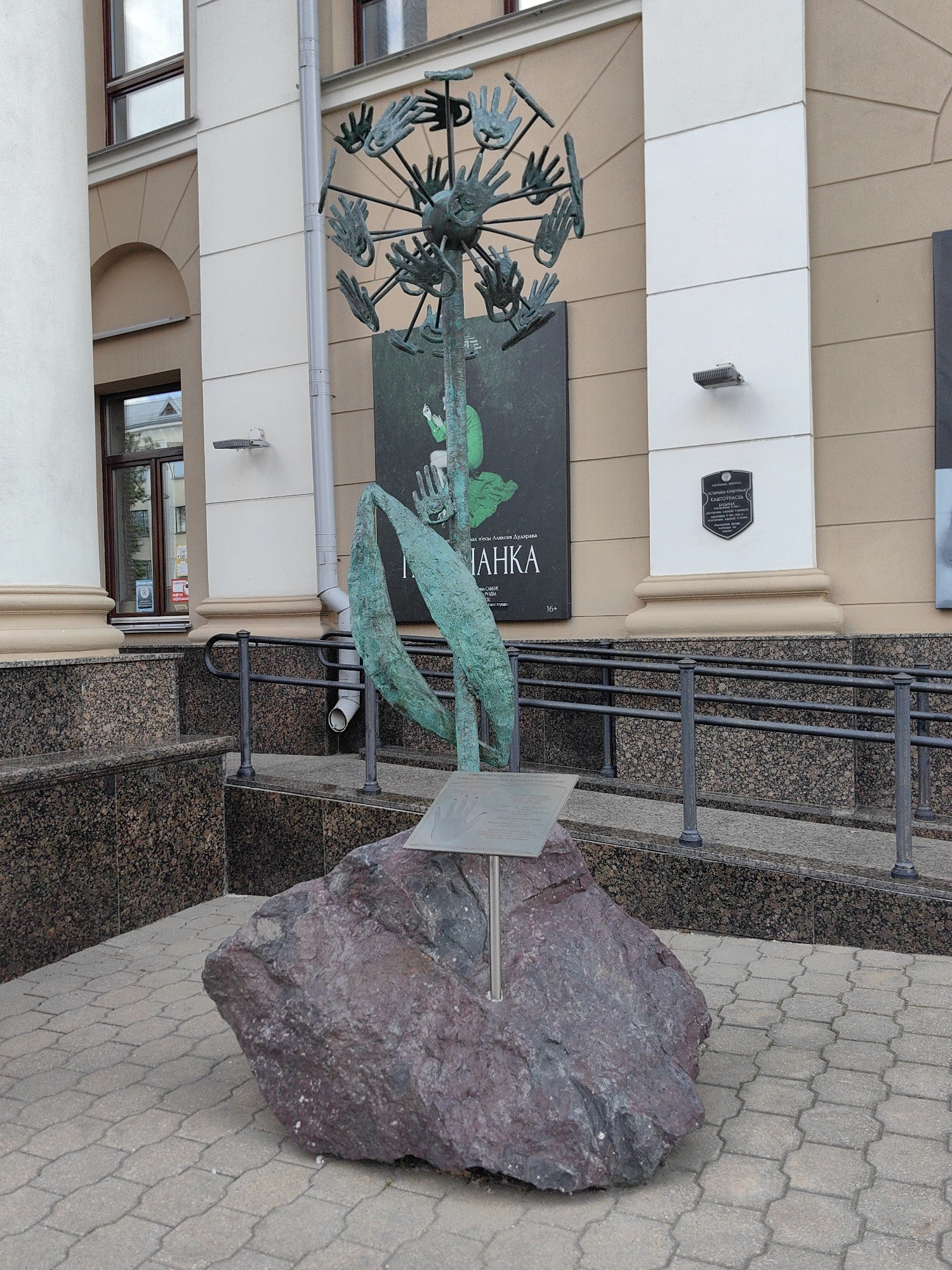The image shows an outdoor sculpture located in front of a building in Minsk, Belarus. The sculpture is abstract, resembling a dandelion with many metal hands forming the seed head. It is mounted on a large, reddish-purple rock, which is situated on a paved walkway. Behind the sculpture, a building with cream-colored and beige facade is visible, featuring windows and architectural details. A dark gray railing runs along the edge of the walkway. A dark green poster with Cyrillic text is mounted on the building behind the sculpture. The overall scene depicts a public art installation in an urban environment.
