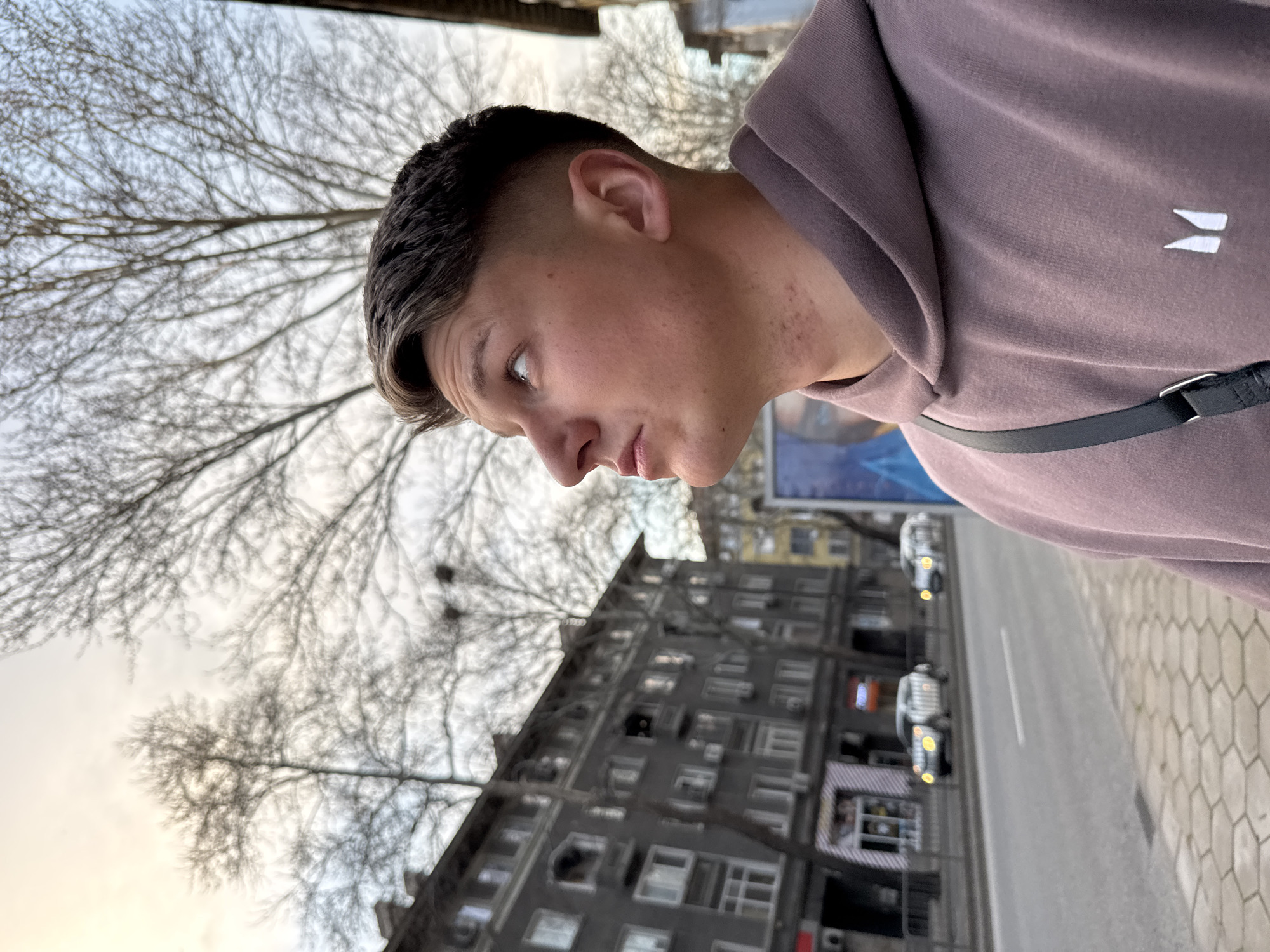 A medium shot depicts a young man in profile, looking down and to the left, wearing a mauve-colored hooded sweatshirt with a white design on the chest, and a black strap over his shoulder. The background features a multi-story apartment building with numerous windows, typical of urban residential architecture. Above the building are bare tree branches against a pale sky. The street and sidewalk are visible, with cars and a shop front with striped awnings suggesting an urban setting. The image is likely taken in Plovdiv, Bulgaria due to the architectural style and general atmosphere consistent with that location.