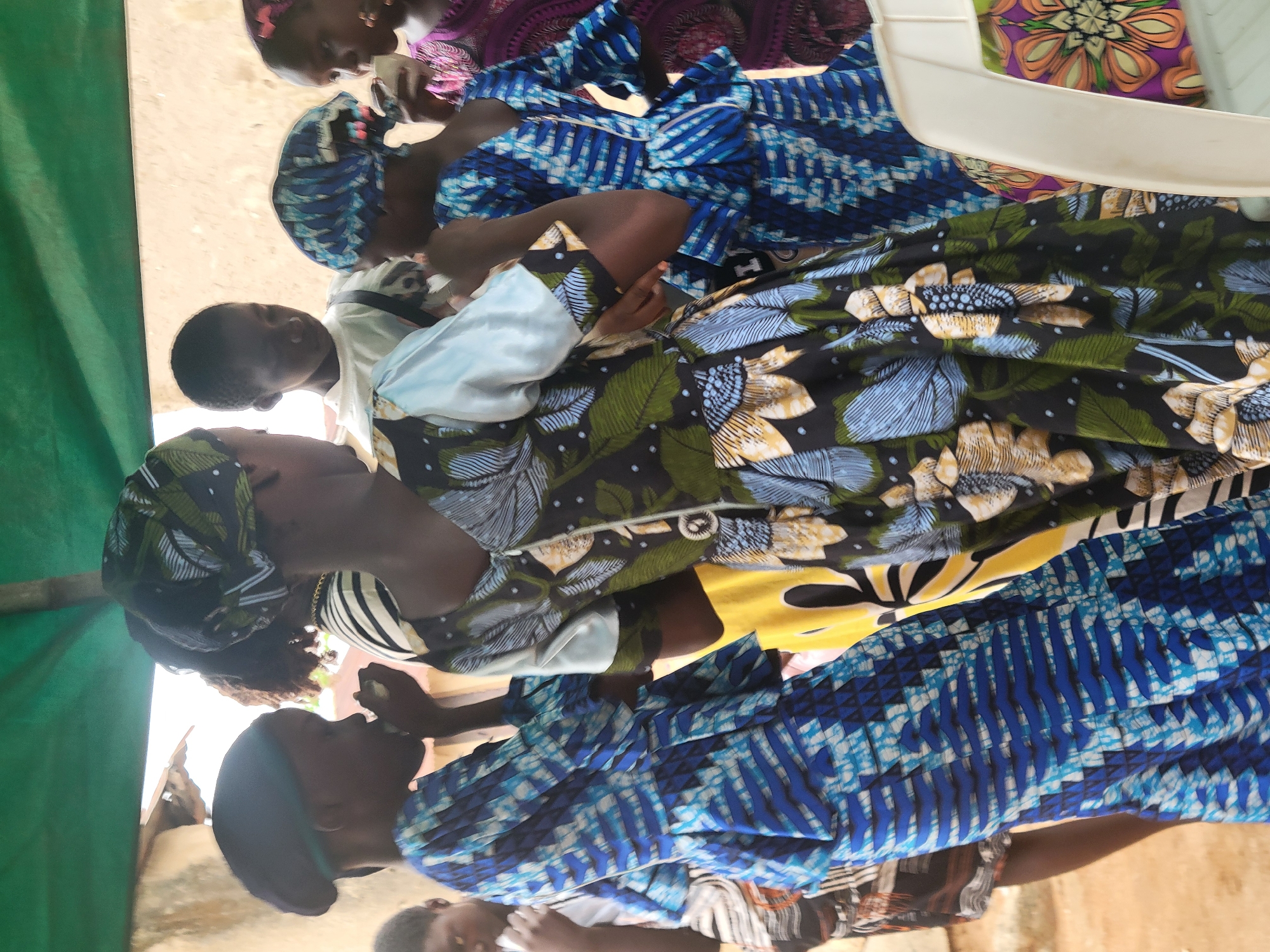 The image shows several people gathered in an outdoor setting in Madalla, Nigeria. The location appears to be under a covered area with a green sheet visible. Several women are present, dressed in colorful, patterned fabrics, including blue tie-dye and floral designs. Some appear to be eating or holding food. There's a white plastic chair partially visible. A child is also present among the people. The environment looks somewhat casual and informal.