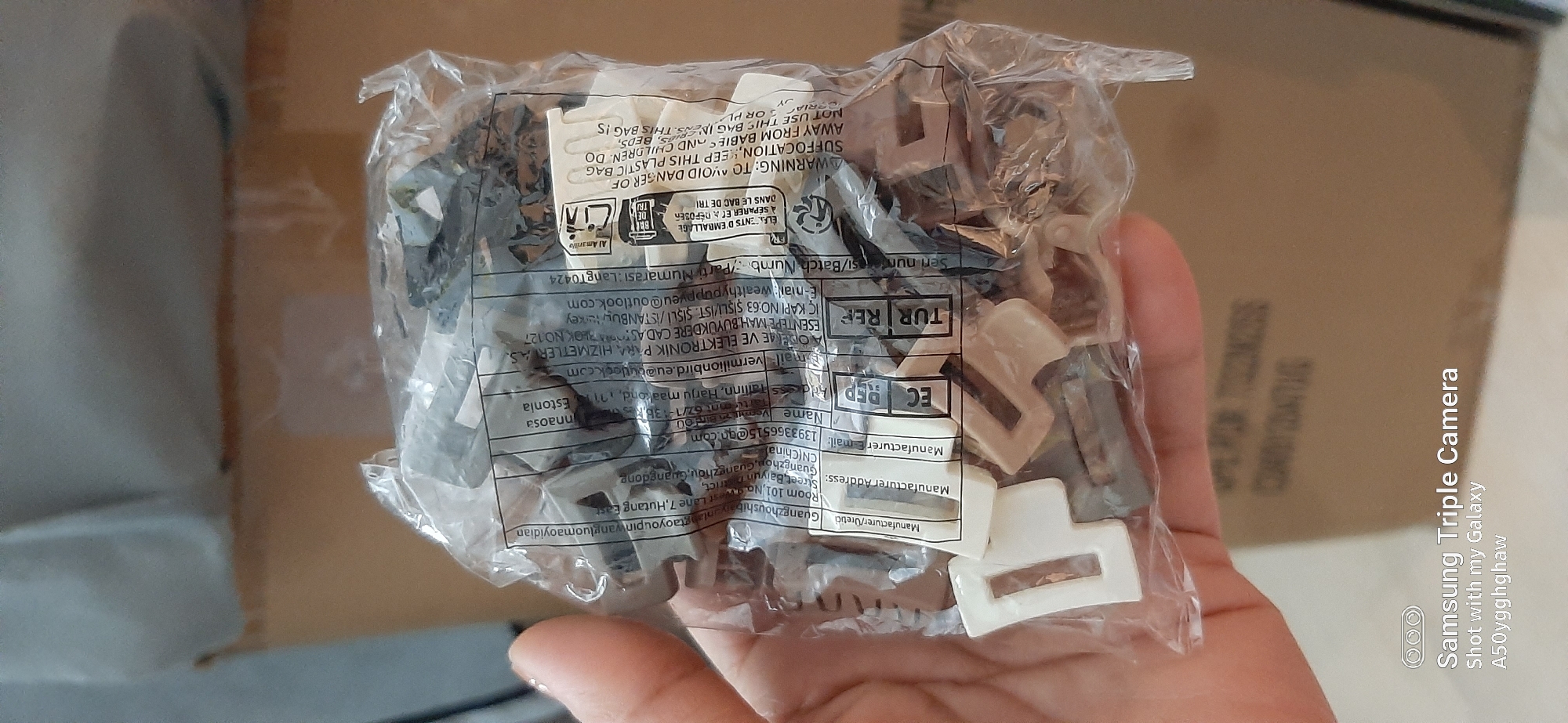 A close-up, top-down shot shows a hand holding a clear plastic bag filled with small, beige and gray plastic clips. The bag has printed labels with text in multiple languages, including English, Turkish, and Chinese. Some of the English text includes "Manufacturer Address," "Manufacturer Email," "EC REP," "TUR REP," "WARNING: TO AVOID DANGER OF SUFFOCATION," and "KEEP THIS PLASTIC BAG AWAY FROM BABIES AND CHILDREN." In the lower left corner, a Samsung Triple Camera watermark is visible. The background is a blurred cardboard box. The image does not provide any information to determine its location in Dubai, United Arab Emirates.