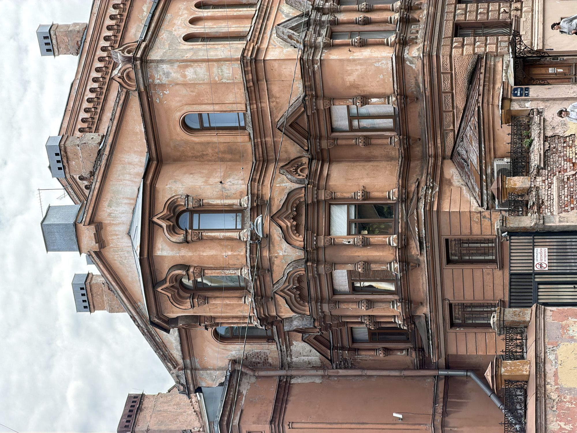An upward-angled view captures the ornate and weathered facade of a multi-story building in an urban setting, typical of Saint Petersburg. The building, rendered in a peach or light terracotta hue, exhibits significant signs of age, with peeling paint, faded plasterwork, and some exposed brick revealing its underlying structure.
Architecturally, the building is highly decorative, featuring a blend of styles with numerous embellishments. Its facade is punctuated by various windows, including arched and rectangular shapes, many adorned with elaborate moldings, decorative columns, and cornices. Several bay window-like projections extend from the building. The roofline, visible against an overcast sky, includes multiple chimneys, some fitted with modern metal caps.
On the right side of the image, two indistinct human figures are visible, seemingly standing near the building, possibly at street level or on a balcony. This section also reveals wrought iron railings, a metal drainpipe, and an outdoor air conditioning unit. Electrical wires or communication cables are strung across parts of the facade.
The scene takes place during the daytime under an overcast sky, which casts a soft, diffuse light. Visible text includes a black and white sign displaying a no-parking symbol with the Russian phrases: "СТОЯНКА АВТОМОБИЛЕЙ ВОСПРЕЩЕНА" (Parking of cars is forbidden) and "НЕ ПАРКОВАТЬСЯ" (Do not park). A smaller sign above it reads "РЫНКОВ О.А. 13," likely indicating a name and number.