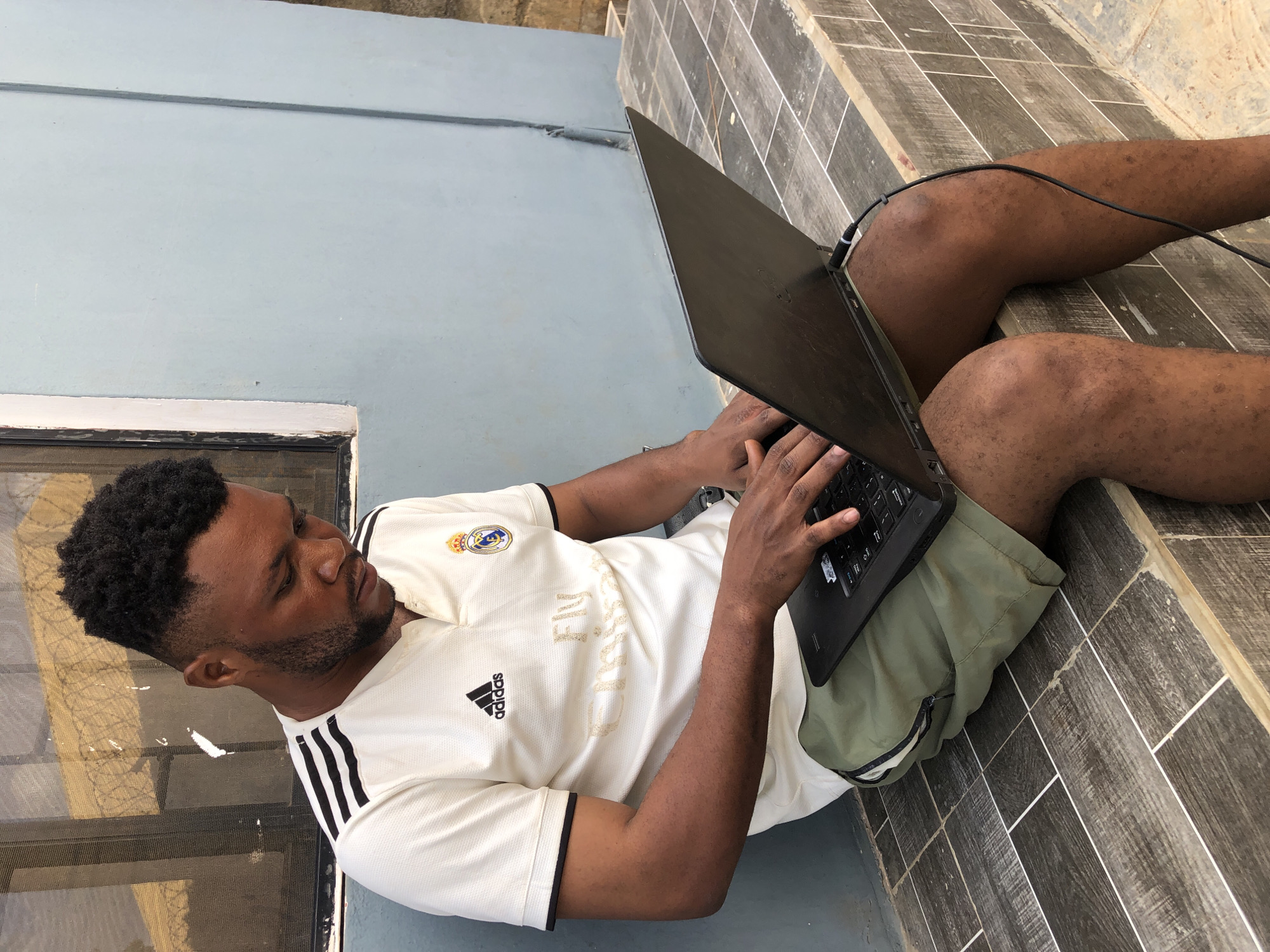A man is sitting on tile steps while using a laptop computer in an unknown city and state. He is wearing a white t-shirt with the Adidas logo and three black stripes on the shoulders, and green shorts. The laptop is black with a power cable. The steps are dark brown with light brown grout. A light blue wall is behind the man.
