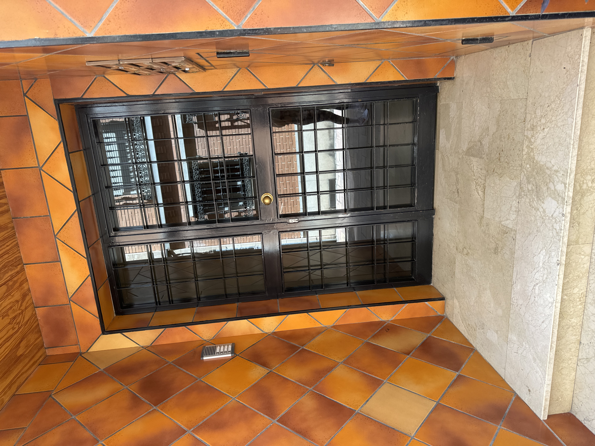 The image shows an entrance to a building with a double door. The doors are made of black metal with glass panels and a grid pattern. The walls on either side of the entrance are tiled with orange and yellow diamond-shaped tiles. The floor is made of light-colored marble tiles, and there are two steps leading up to the entrance. A person is reflected in the glass doors. The location is a street in the city of Murcia, Spain.