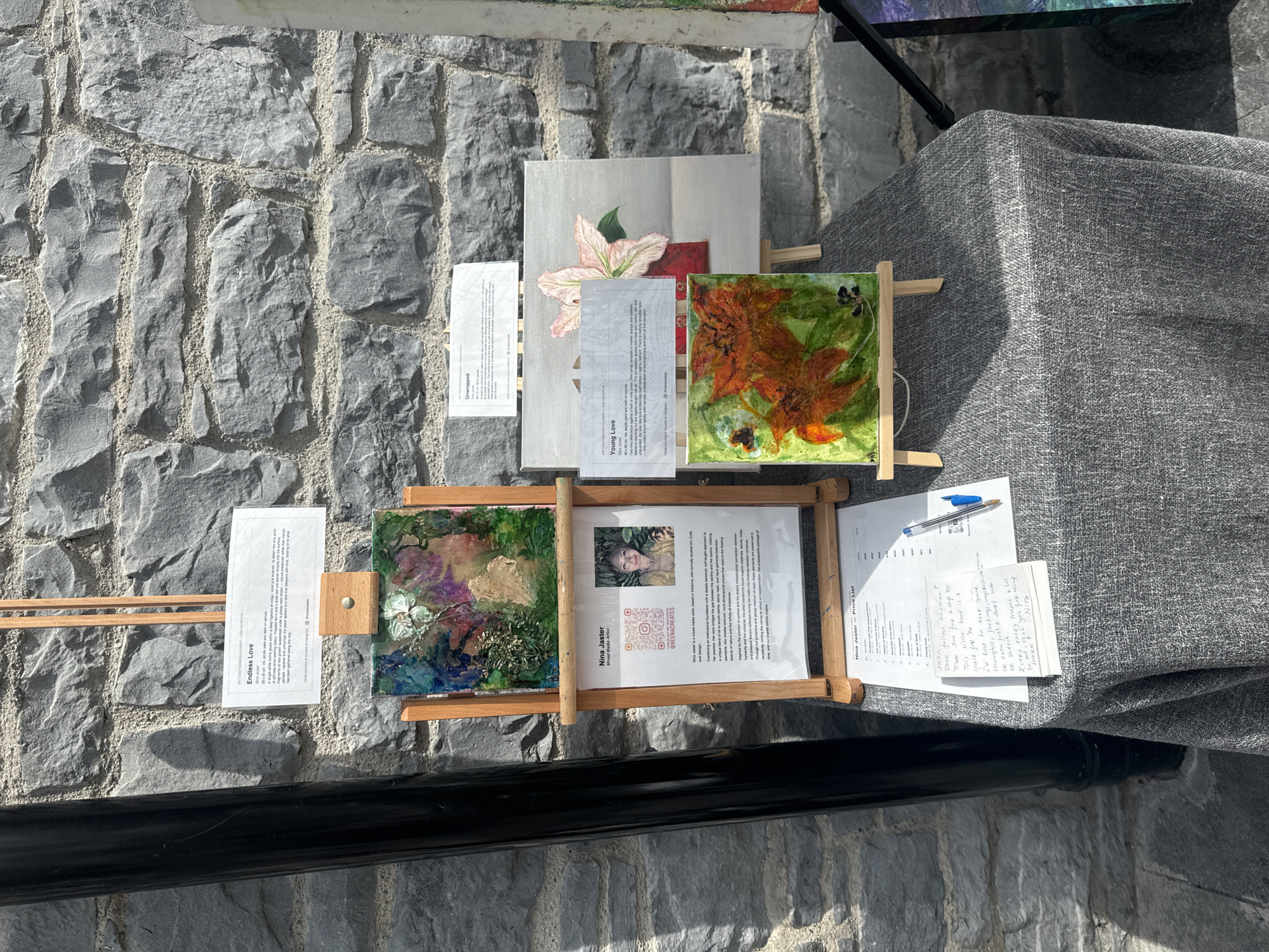 This image depicts an art display in the city of Kilkenny, Ireland, likely outdoors given the stonework and natural lighting. The primary focus is on several artworks presented on wooden easels and displayed on a grey linen-covered table.
In the foreground, a small, colorful painting with orange and green hues, possibly featuring abstract floral elements and a butterfly, is propped on a miniature easel. Next to it, a larger canvas displays a delicate pink lily in a red vase. A descriptive card with the title "Young Love" and the artist's name, Nina Jaster, is placed in front of this painting.
Further back, a more intricate mixed-media artwork titled "Endless Love" by Nina Jaster is displayed on a larger easel. This piece features a textured surface with blues, purples, and green elements. Accompanying this artwork is a more detailed information sheet about Nina Jaster, a mixed-media artist from Kilkenny, which includes her photograph, QR codes, and a description of her artistic approach. Below this, a price list for Nina Jaster's works is visible, along with a handwritten note from the artist and a blue pen.
The backdrop consists of a rough-hewn stone wall, characteristic of historic architecture. The overall scene suggests an art exhibition or market stall, showcasing local art in a public space in Kilkenny.