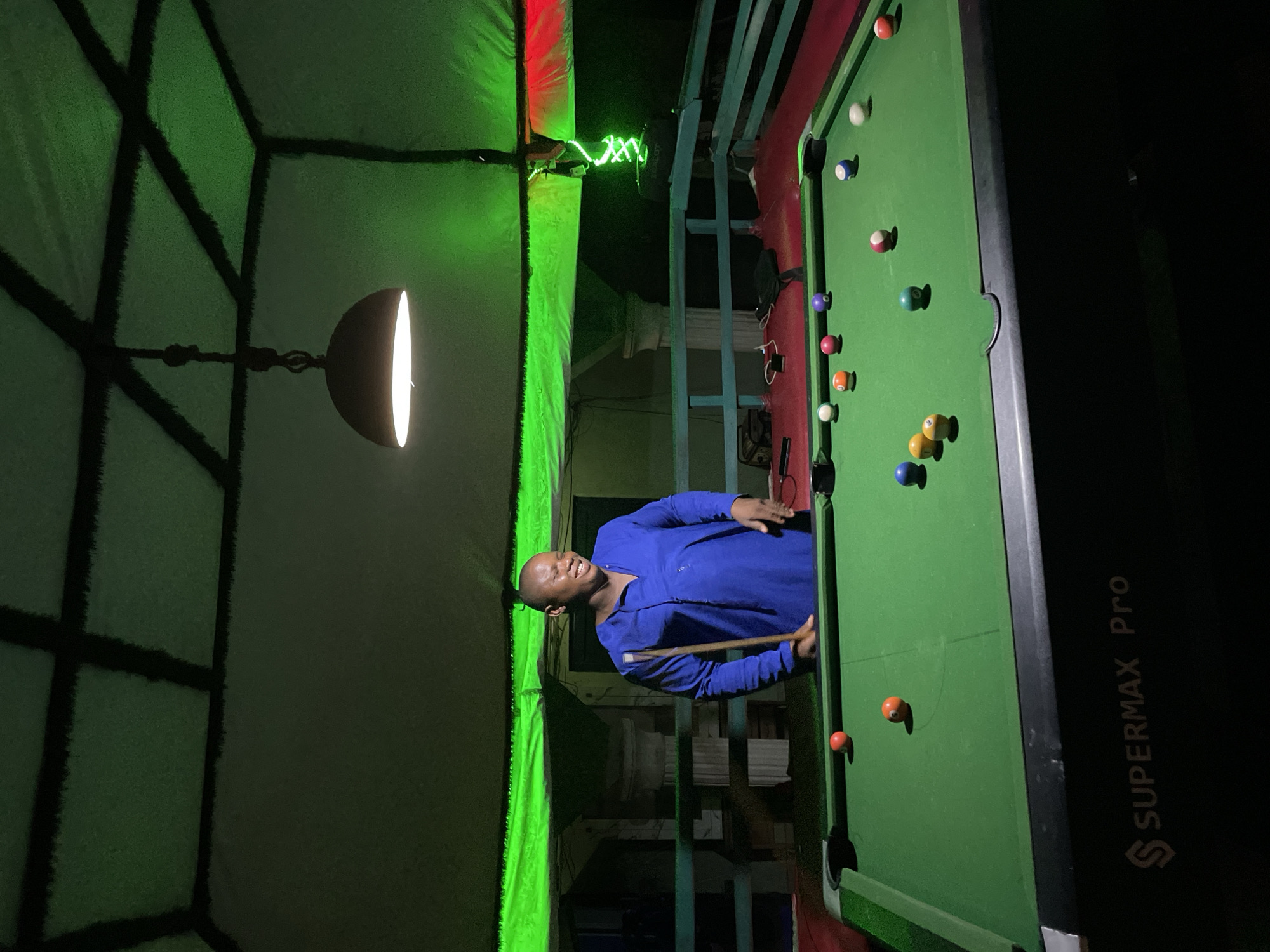 The image depicts a recreational scene inside an establishment in Lokoja, Nigeria, at night.
A dark-skinned adult male with a bald head stands on the left side of a green-felted pool table. He is wearing a long-sleeved, royal blue tunic-style shirt and is smiling or laughing, looking towards the right side of the frame. He holds a wooden pool cue in his right hand, resting it on the table's edge.
The pool table, branded "SSUPERMAX Pro" on its side, is central to the image. Several multi-colored pool balls are scattered across its green surface, including a white cue ball, solid red, solid orange, solid yellow, solid green, solid blue, solid purple, solid pink/magenta balls, and striped orange and red/pink balls.
Above the table, a single, downward-facing, bowl-shaped pendant lamp provides localized white illumination. The ceiling features a light-colored, possibly fabric or canvas, covering with dark intersecting beams that form square panels. This ceiling and the upper walls are illuminated by green LED strip lighting, with a section of red LED lighting visible on the far right. A spiral-wrapped green light fixture is also visible on the right side.
In the background, behind the man and the pool table, a light blue wooden railing runs horizontally. Beyond the railing, a white fluted pillar or column and stacked wooden crates are discernible. The surrounding area beyond the railing is dark. Objects such as a bag, cables, and a power adapter are visible on the reddish-brown floor or platform area behind the railing.
